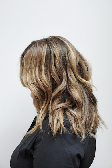 Side view of a woman with wavy, shoulder-length, light brown hair with blonde highlights, wearing a black top against a plain white background.
