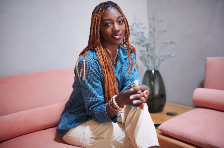 A woman with long braided hair, wearing a blue checkered shirt and beige pants, sitting on a pink sofa in a modern living room, smiling and looking at the camera.