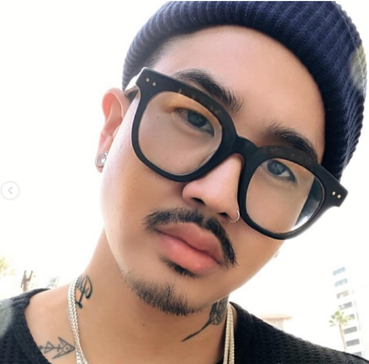Close-up of a young man with black hair, wearing black glasses, a navy beanie, earrings, and necklaces, with tattoos on his neck, outdoors on a bright day.