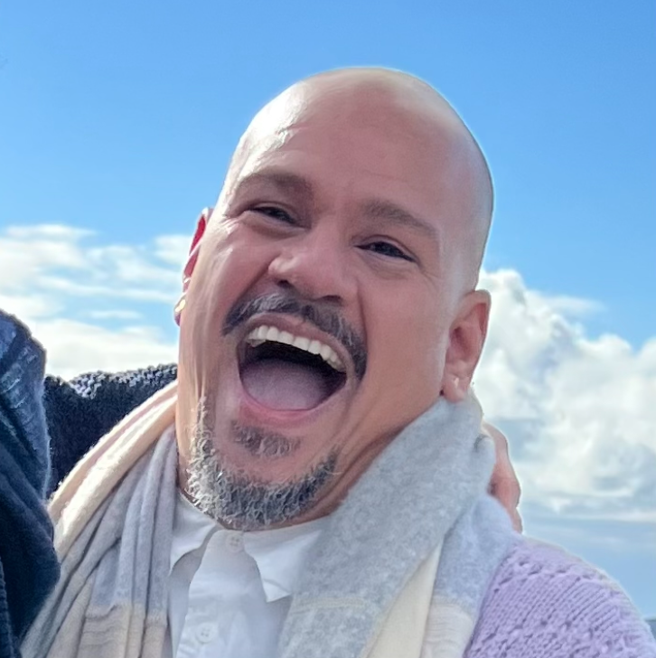 A man smiling and laughing outdoors against a blue sky with clouds, wearing a light-colored scarf and a white shirt.