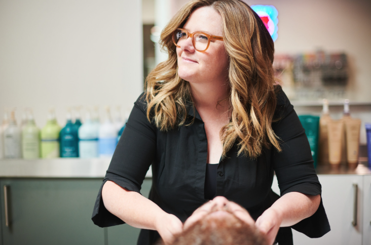 A woman with wavy, shoulder-length hair and glasses, wearing a black shirt, standing in a hair salon with hair care products in the background.