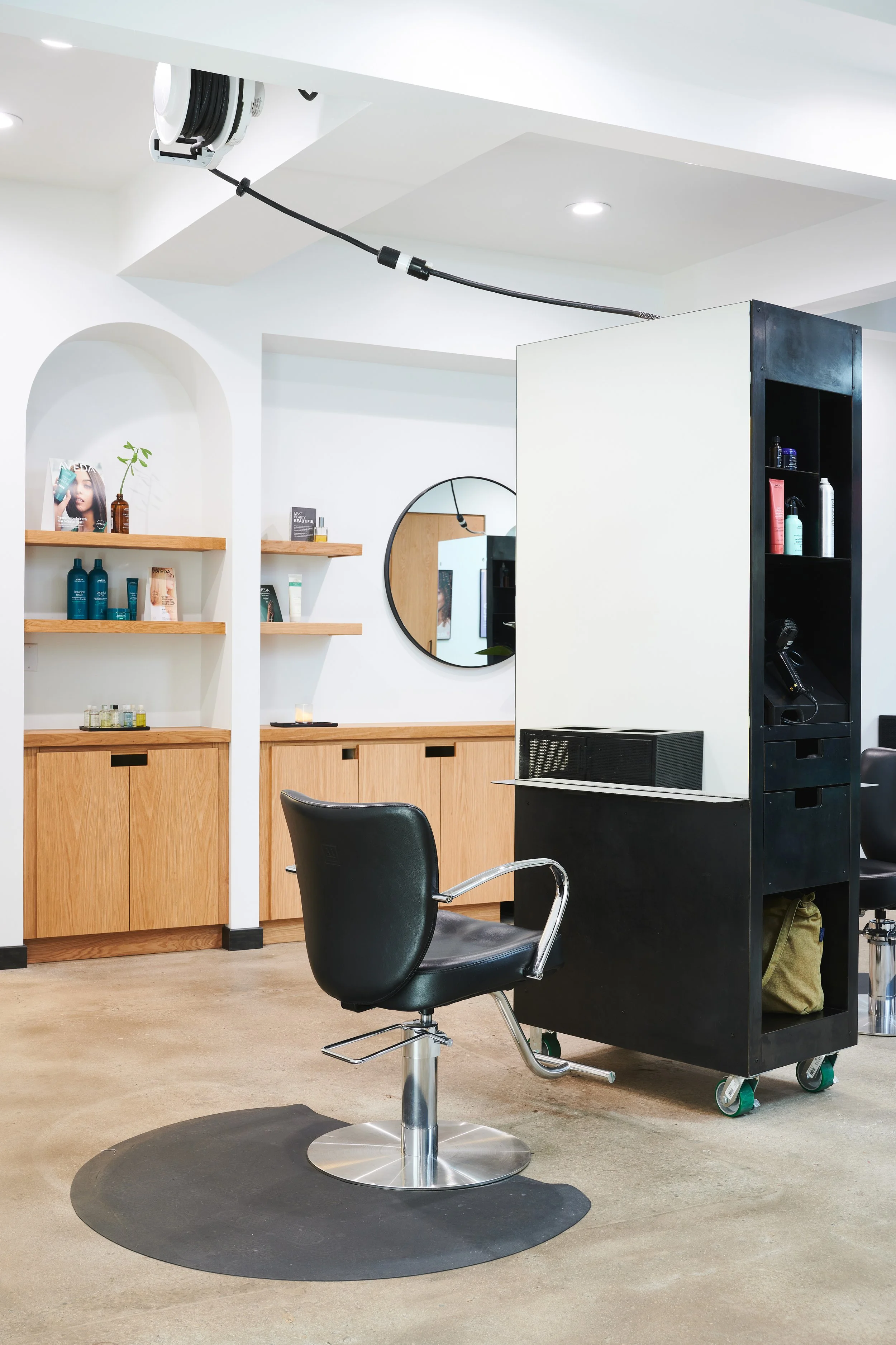 A modern salon interior with barber chairs, wooden shelving displaying hair products, and a black storage unit with more products, a mirror, and a handbag.