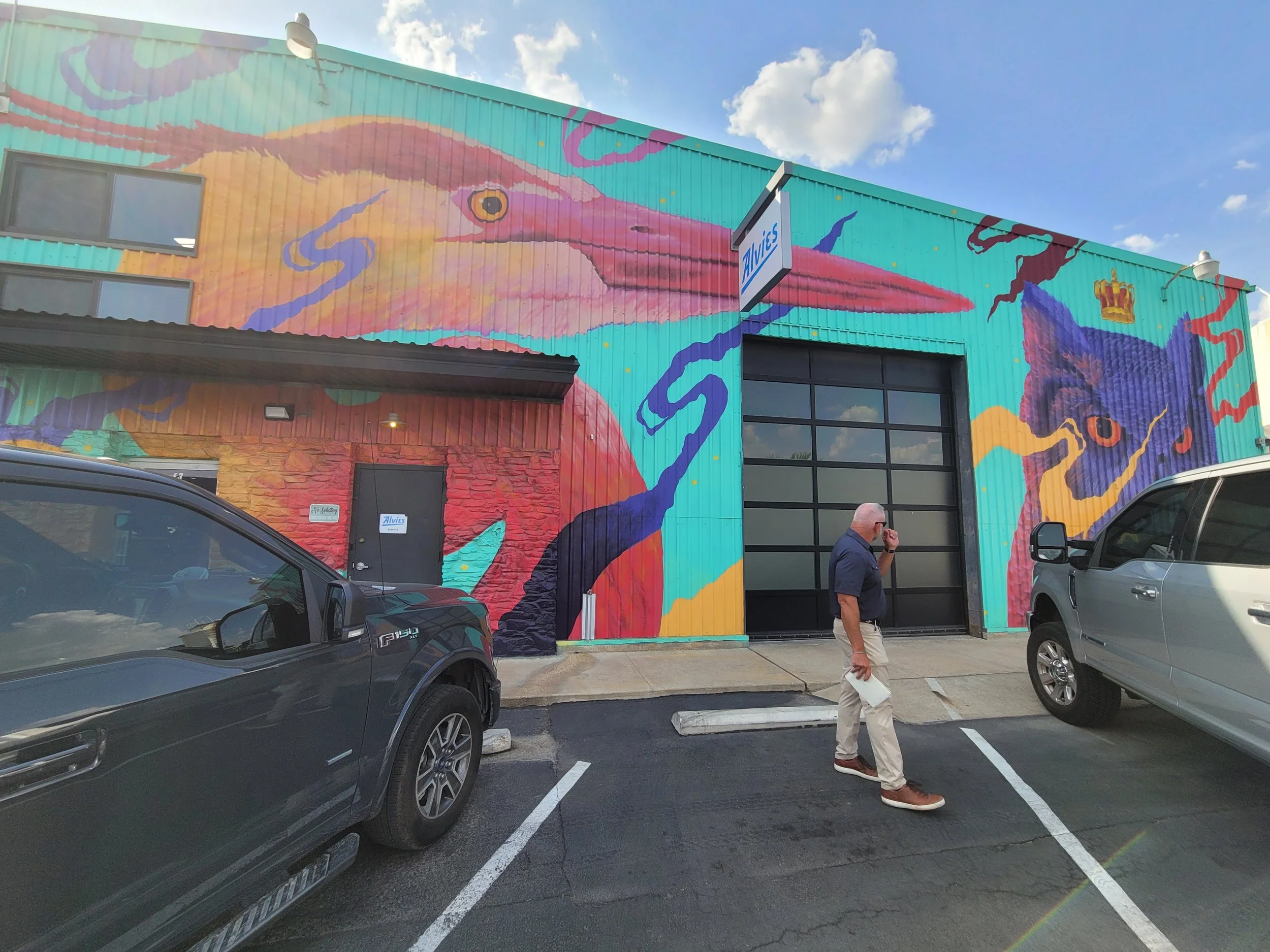 Colorful mural of a bird, possibly a heron or pelican, on the exterior of a building with parking lot and a man walking while talking on his cellphone.