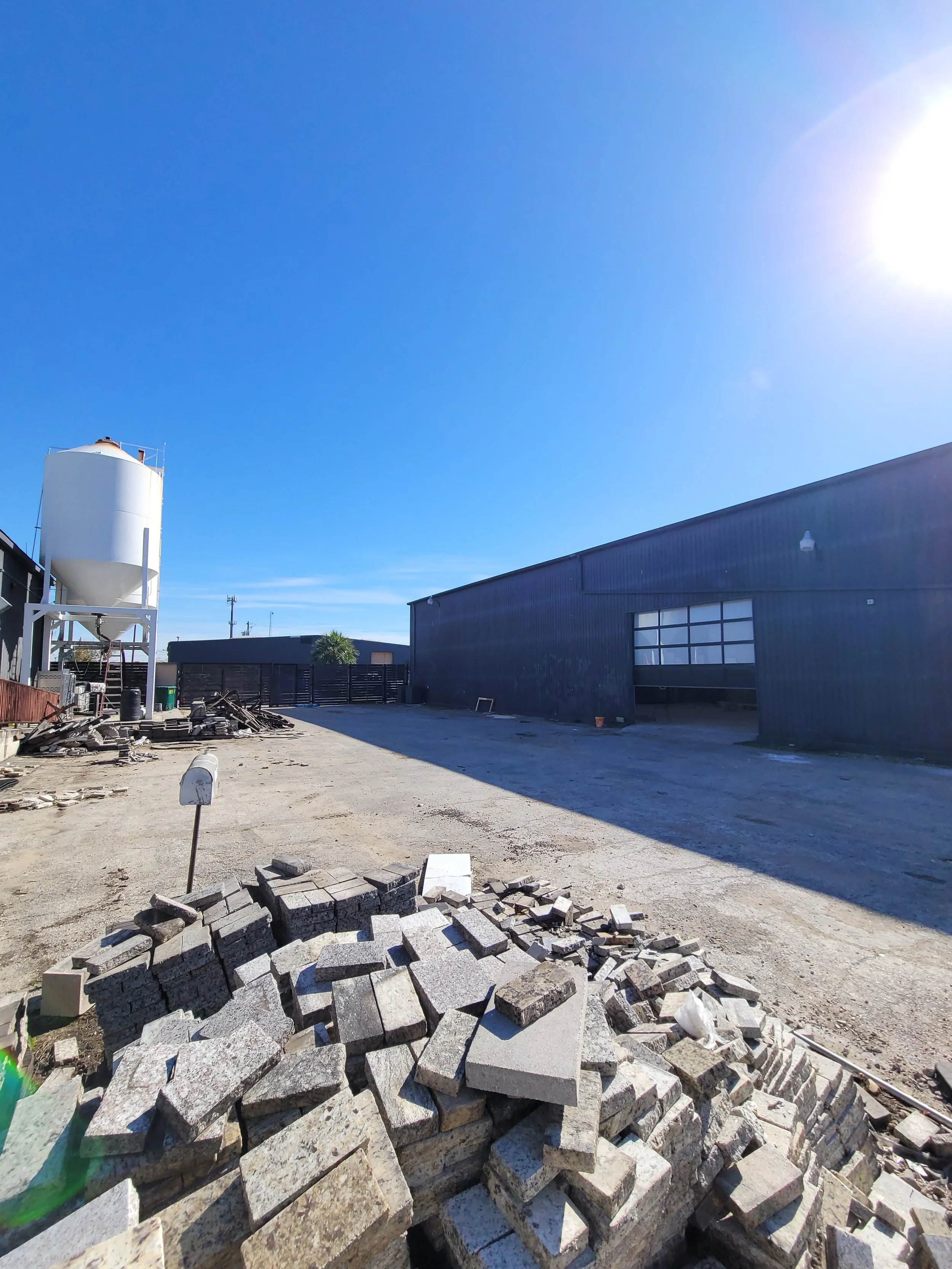 Construction yard with a pile of bricks and concrete blocks in the foreground, a black building on the right, and a silo on the left under a clear blue sky with bright sunlight.