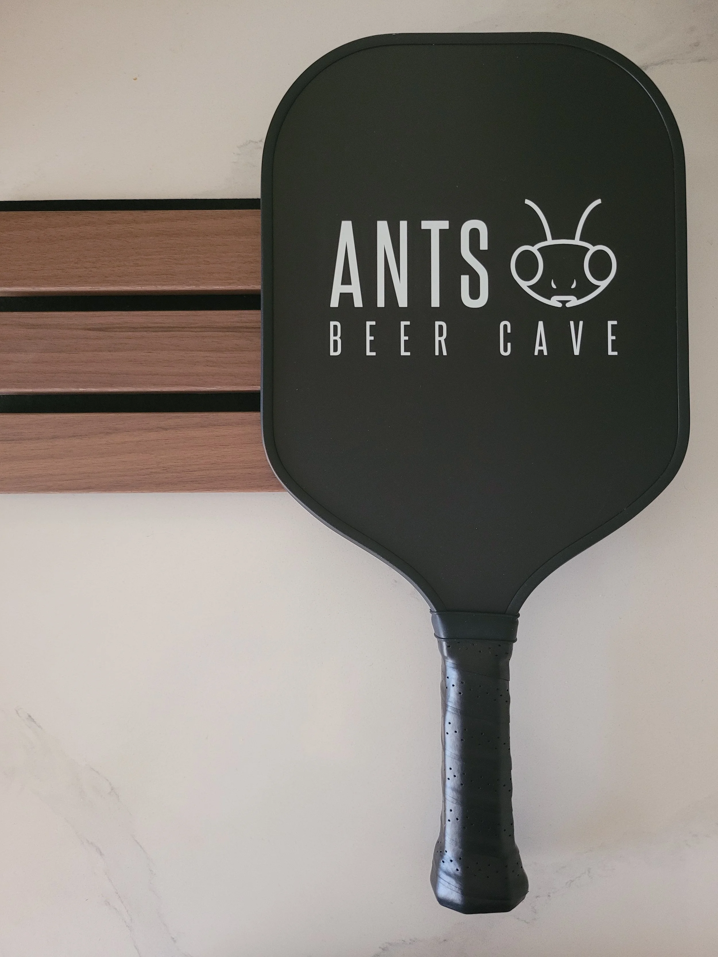 A black paddle with a cartoon ant face and the text 'ANTS Beer Cave' on it, hanging on a wall next to a wooden slat.