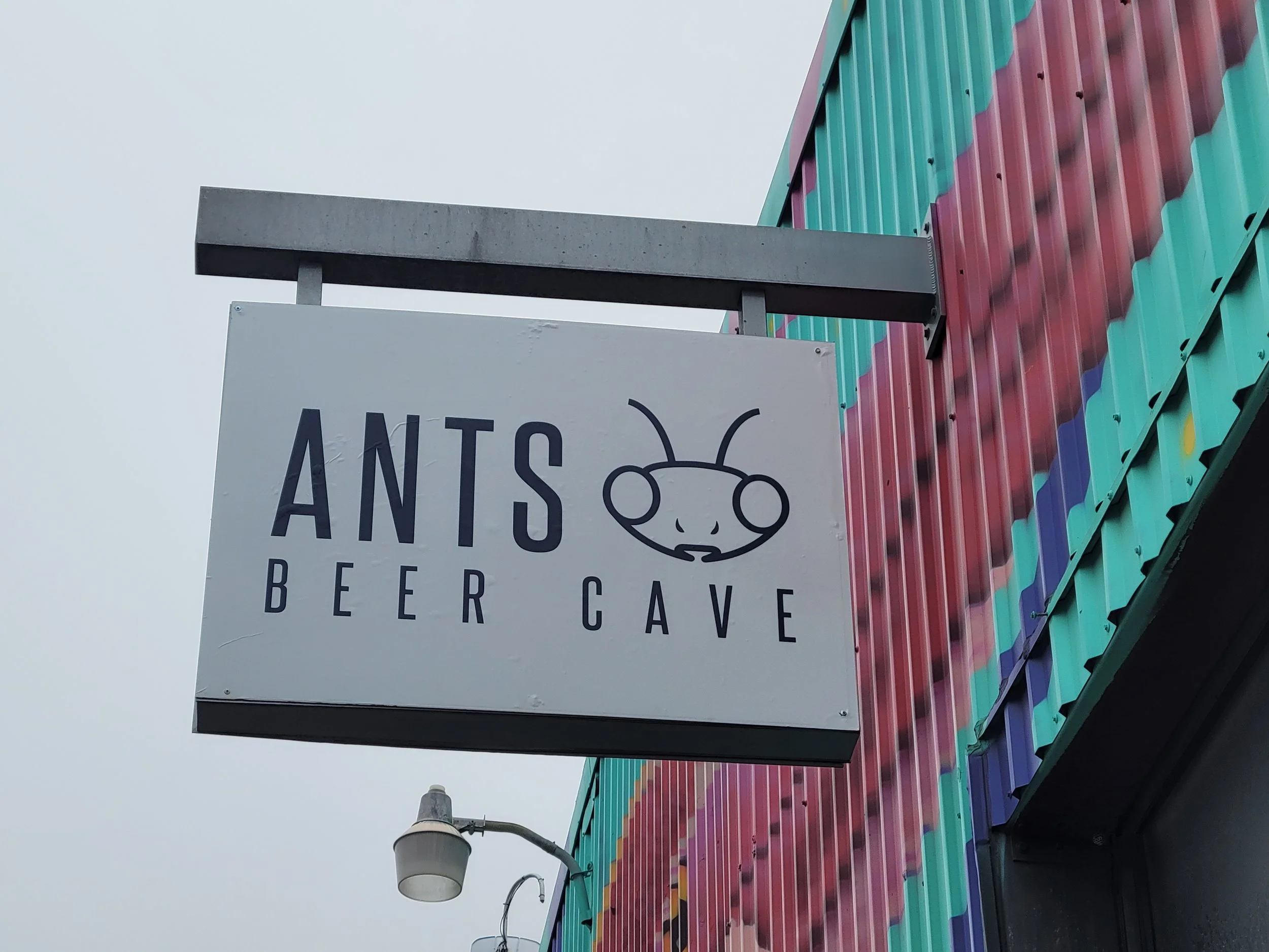 Sign for Ants Beer Cave with a cartoon ant face on it, hanging outside a colorful building.