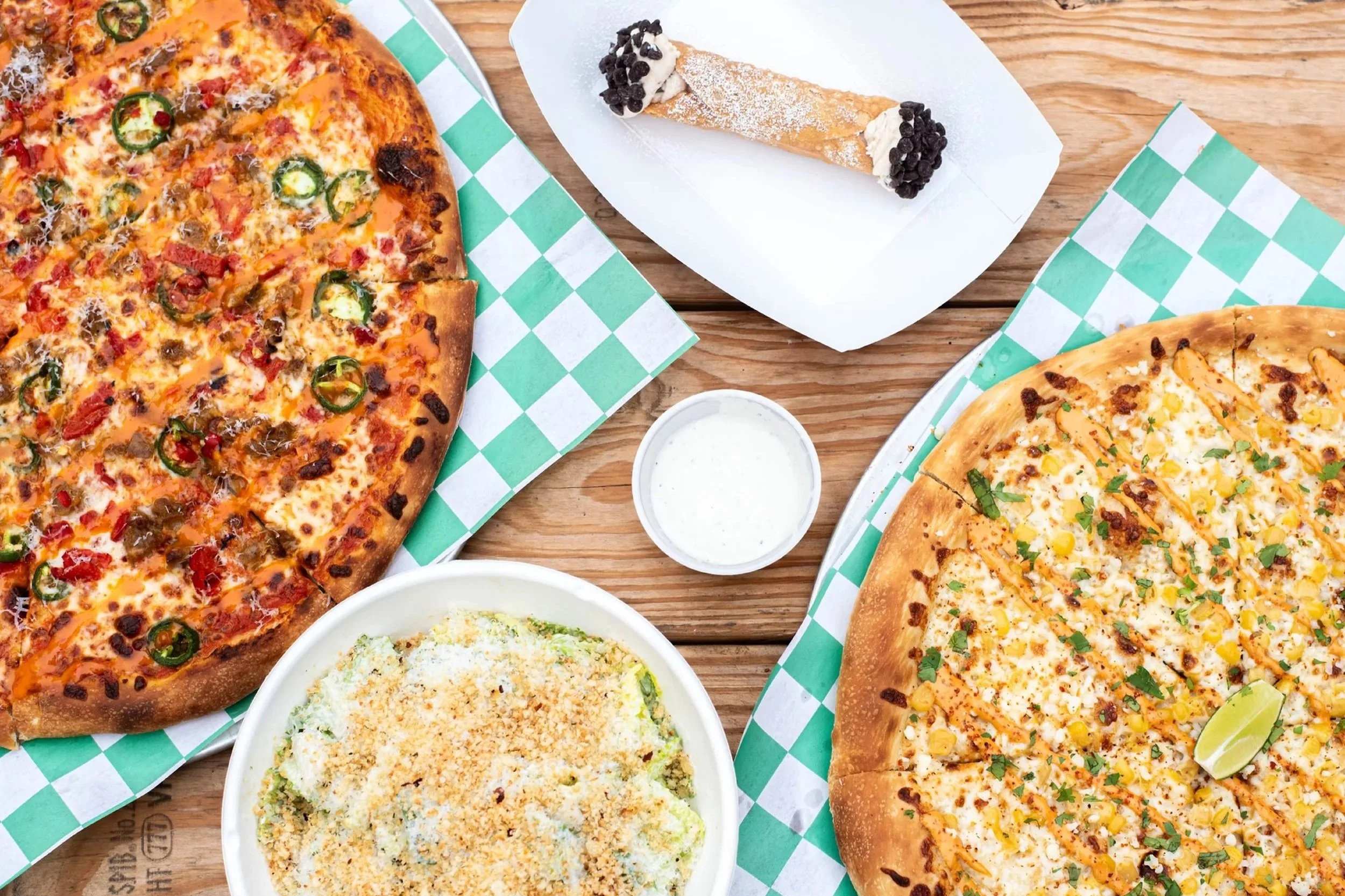 Two pizzas with different toppings, a bowl of salad, a container of ranch dressing, a slice of dessert, and a lime wedge on a wooden table.