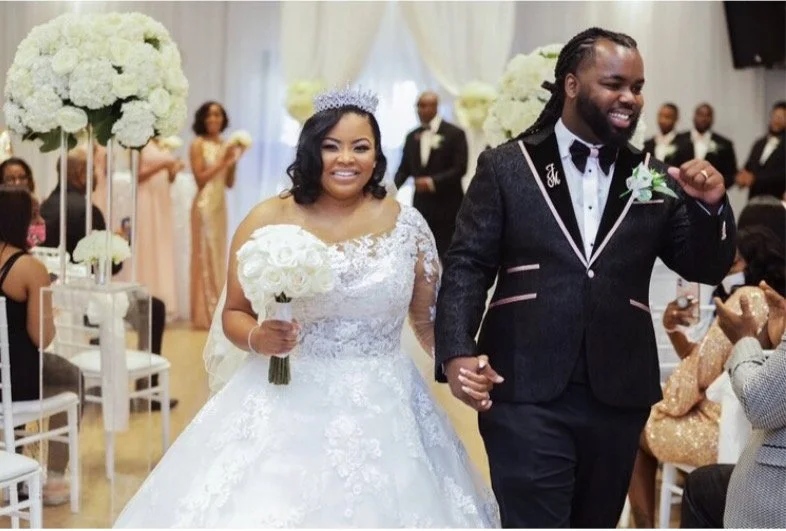 A newly married couple walking down the aisle at their wedding ceremony, smiling and holding hands. The bride is wearing a white wedding gown with lace details and a tiara, holding a bouquet of white roses. The groom is dressed in a dark tuxedo with a bow tie, with a boutonniere, and appears excited. Guests are seated on either side, and there are large white floral arrangements in the background.