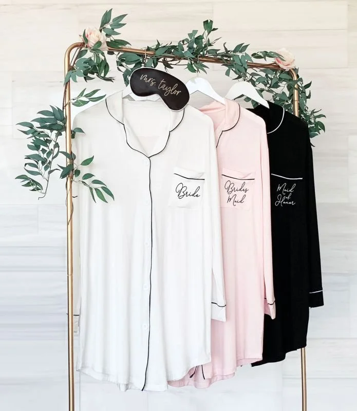 Three satin pajama sets with 'Bride,' 'Bride Maid,' and 'Maid of Honor' embroidered on the chest pockets, hanging on a golden clothing rack decorated with pink roses and greenery, against a white background.
