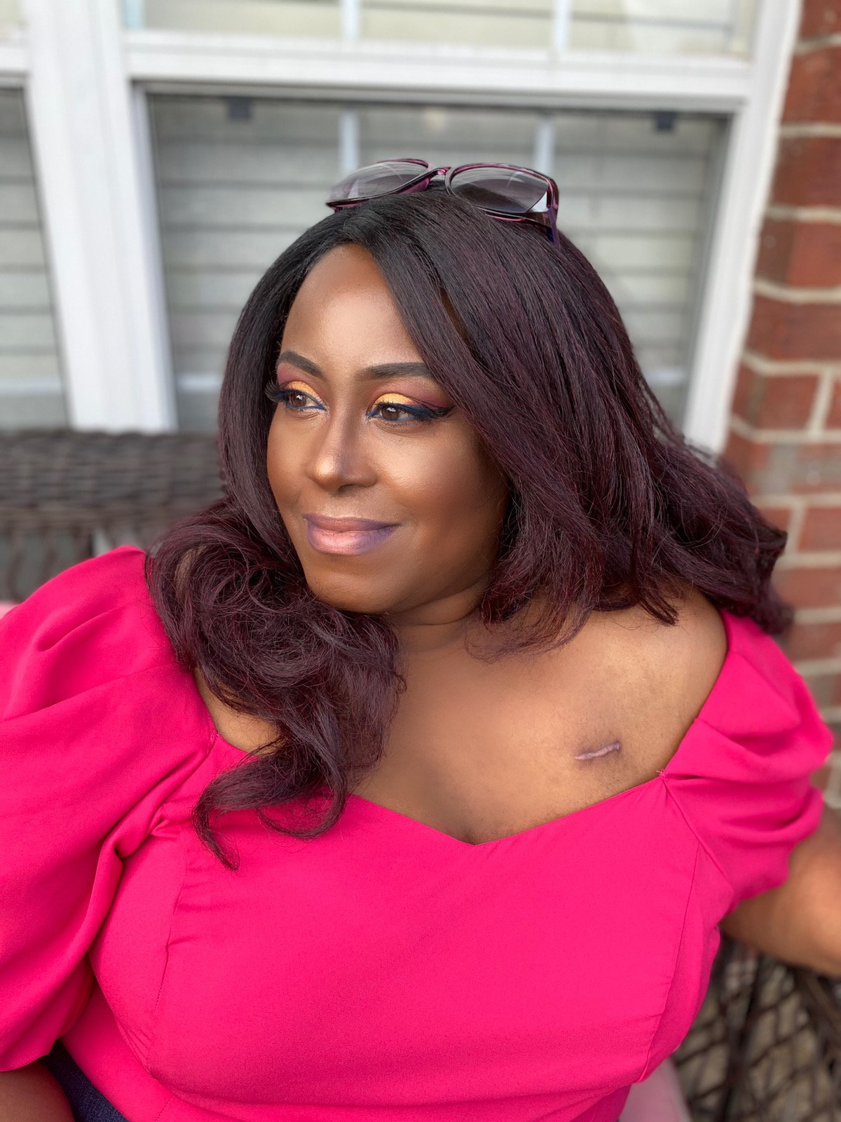 A woman with long, dark hair and glasses on her head, wearing a pink dress, sitting outdoors near a brick wall and window.