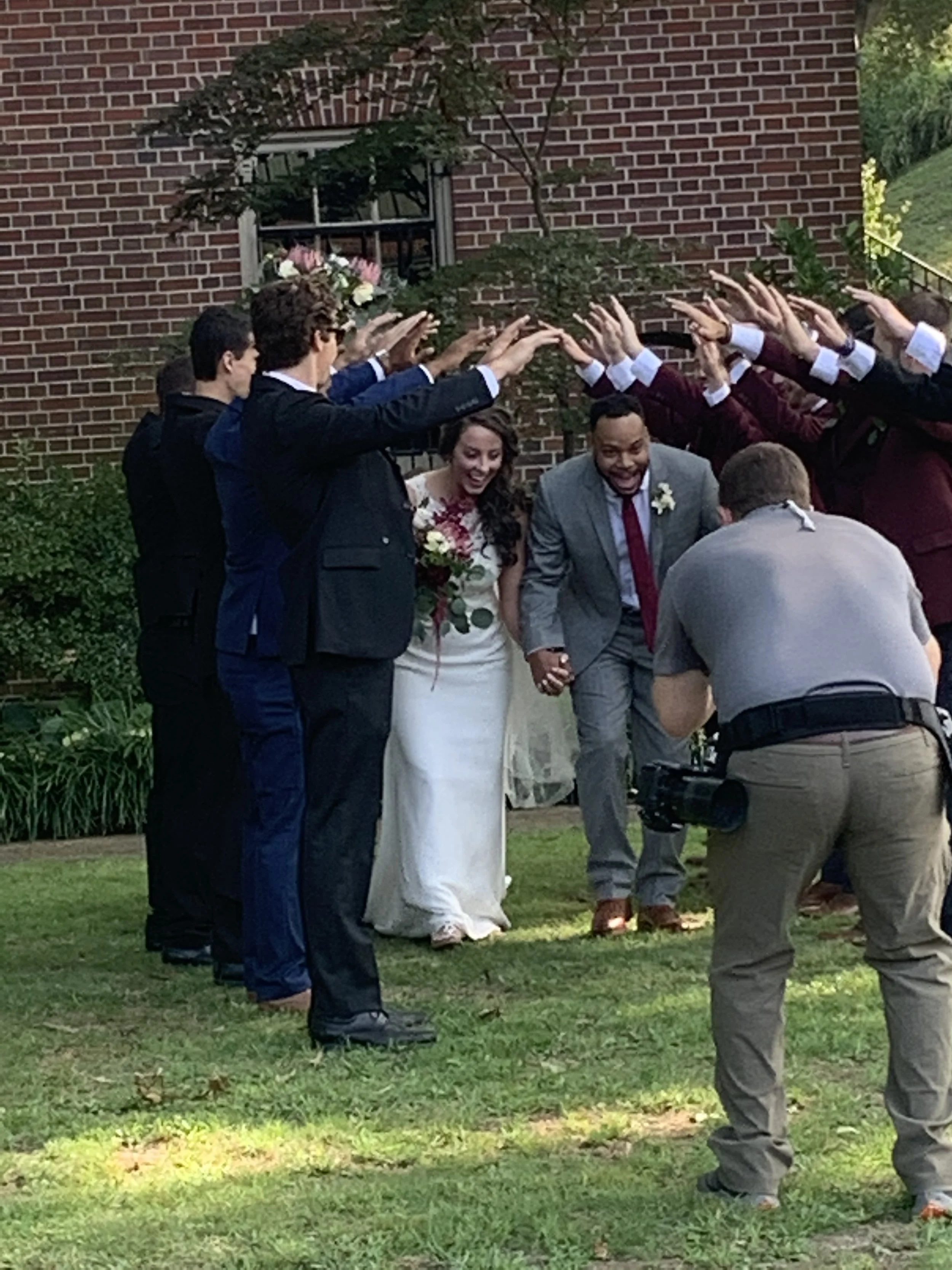 Wedding celebration with bride and groom holding hands, surrounded by friends raising their hands in a pom-pom gesture outside a brick building.