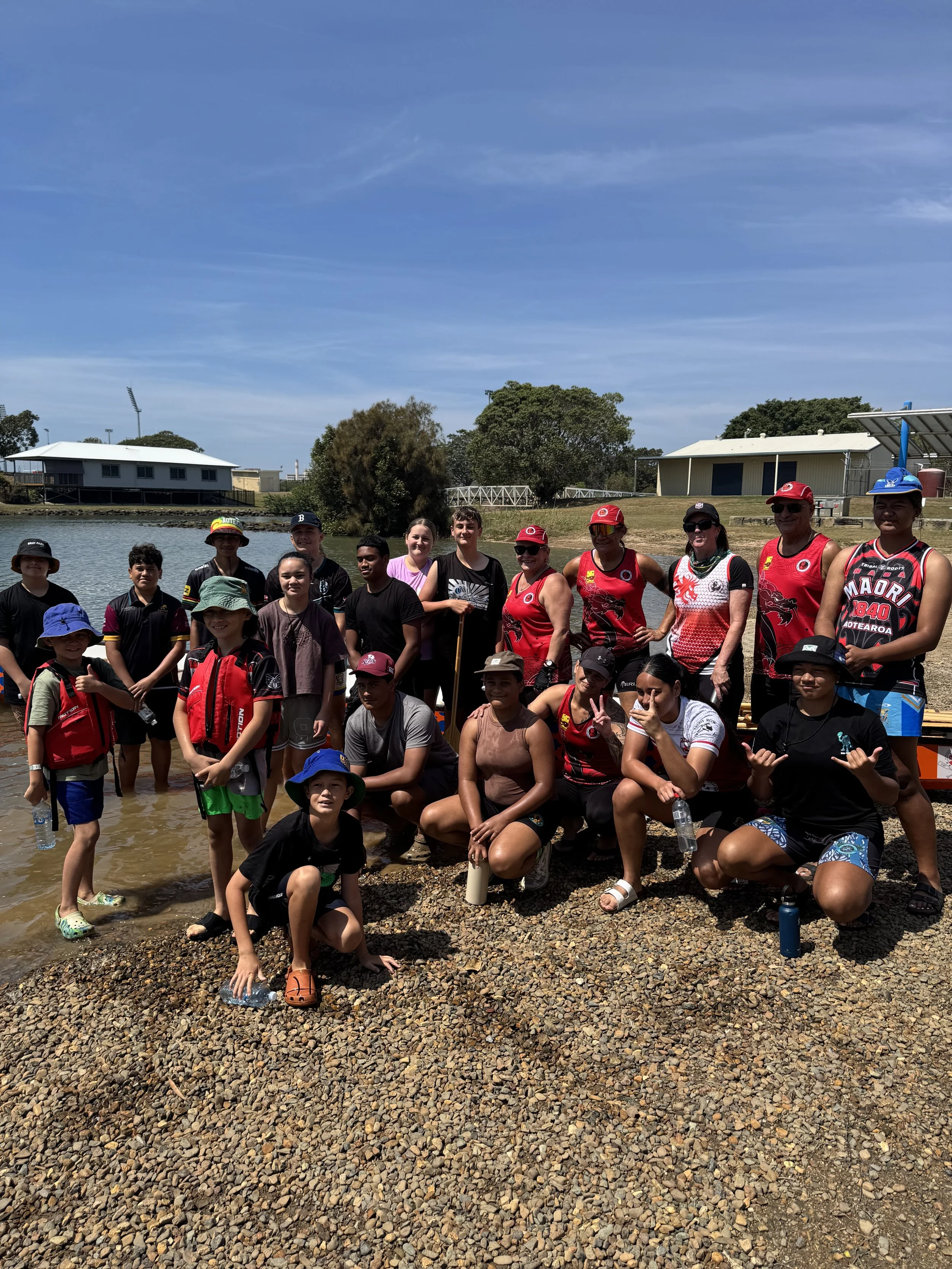 A group of children and adults posing together on a rocky shoreline near a body of water, with trees and buildings in the background, under a clear blue sky during daytime.