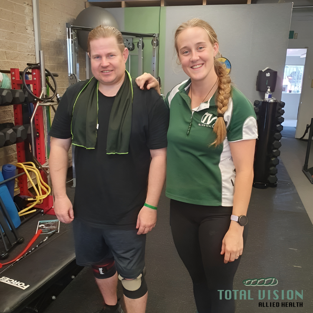 A man and a woman smiling at the camera in a gym. The man has blond hair, is wearing a black shirt, shorts, a knee brace, and a green towel around his neck. The woman has blond hair in a braid, is wearing a green and white sports shirt and black pants. Behind them are gym equipment including weights and resistance bands.