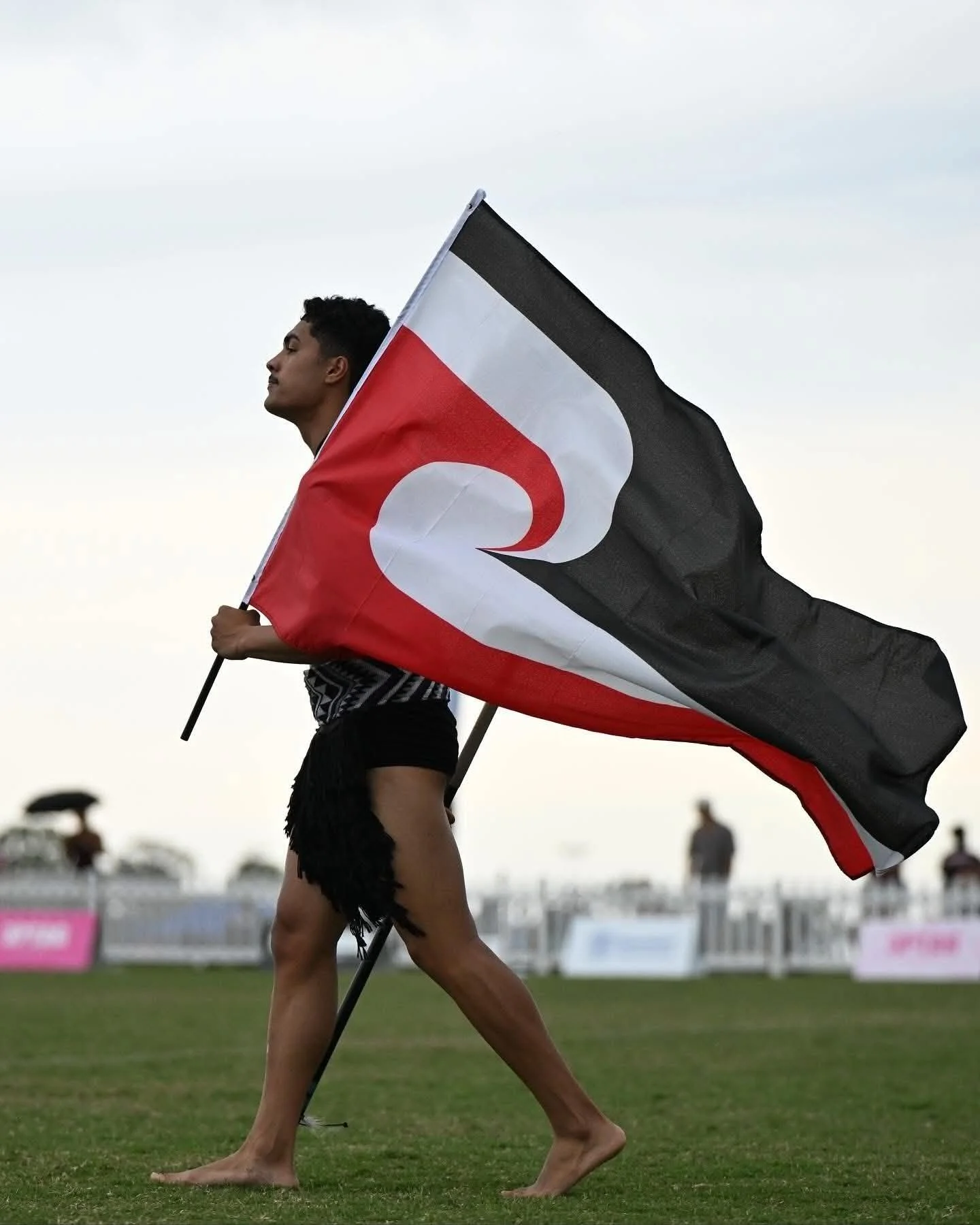 A person holding a red, white, and black flag representing New Zealand Maori Culture, walking barefoot on grass at a sports event.