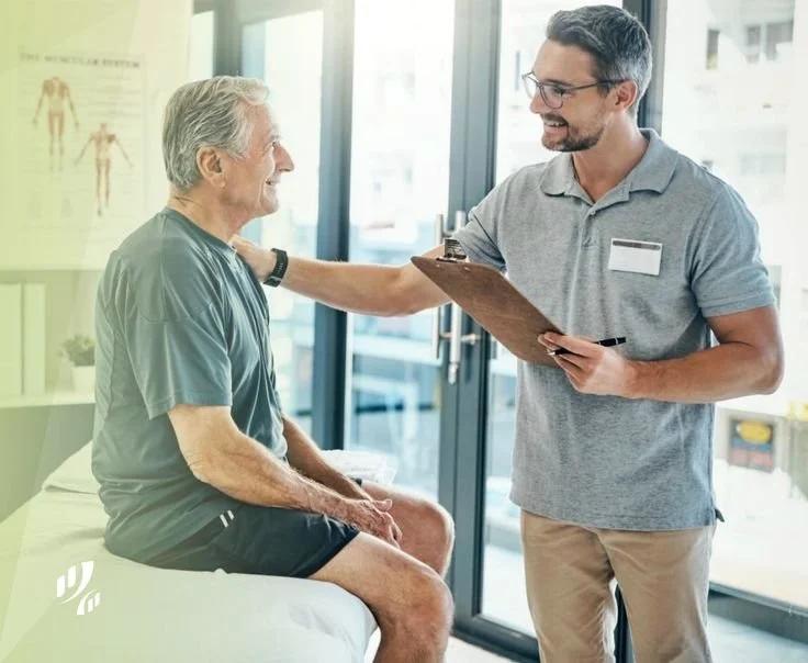 A healthcare professional talking to an elderly male patient sitting on a medical examination table in a clinic or hospital room with large windows.