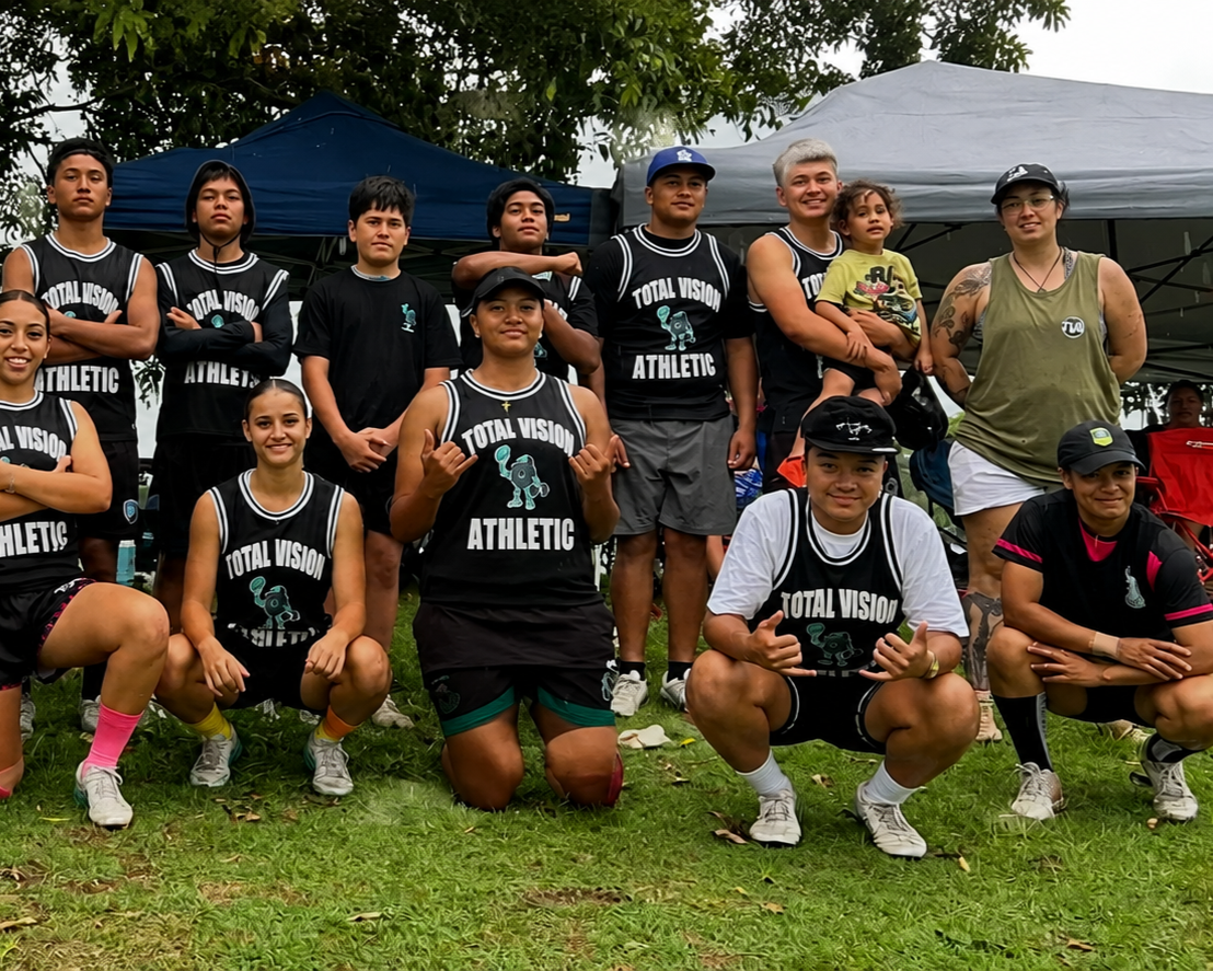 Group of young athletes wearing black jerseys with 'Total Vision Athletic' logo, posing outdoors on grass, with tents and trees in background.