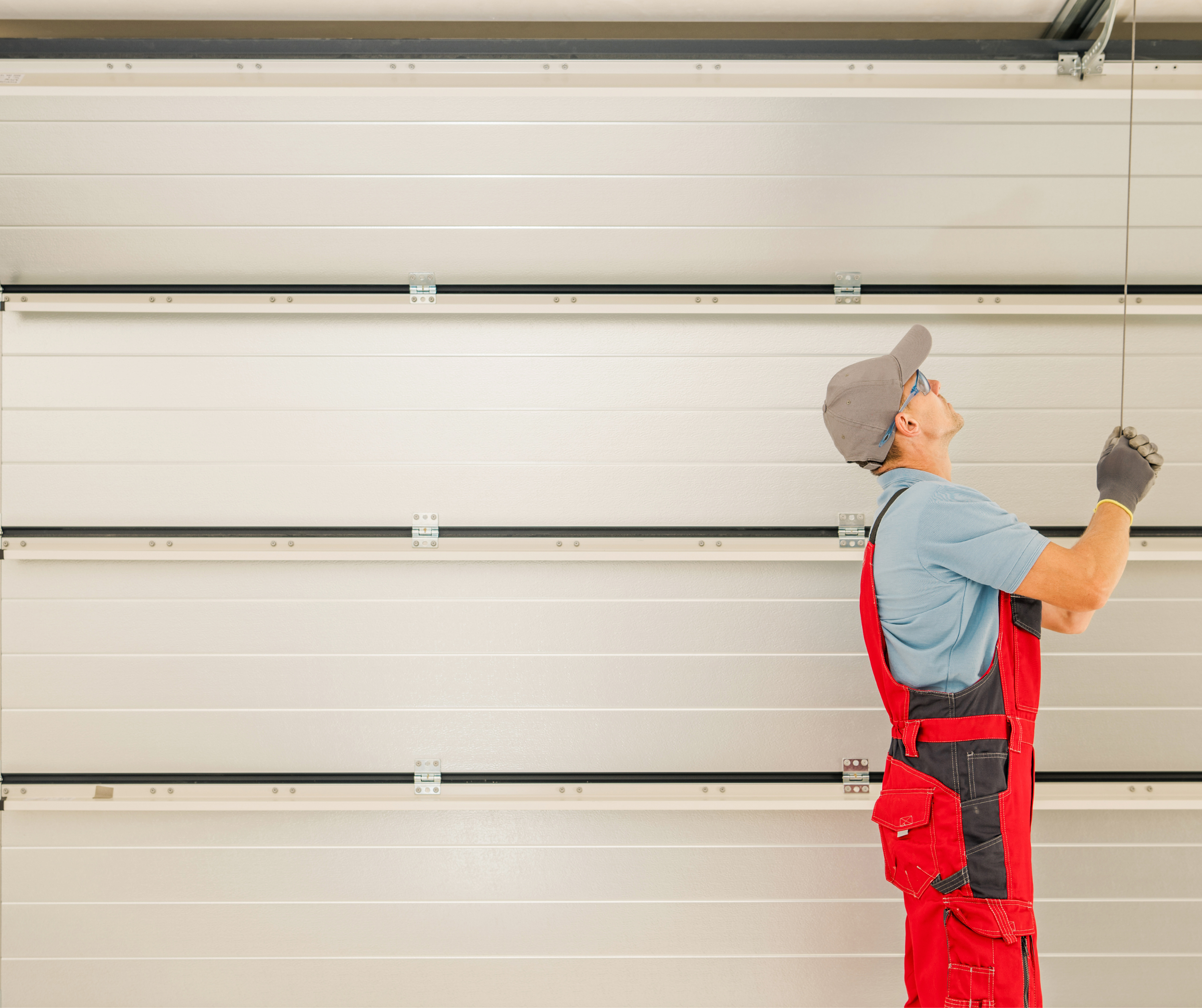 Man in red work overalls and a gray cap standing in front of a large white garage door, looking up and holding a tool in his hand.