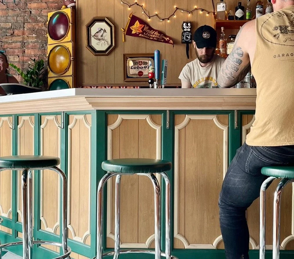 Bar interior with a wooden counter, green and beige decorative panels, two bar stools, a man in a yellow shirt sitting, a bartender wearing a cap and shirt, beer taps, bottles on shelves, framed pictures, a traffic light decoration, a pop art style b