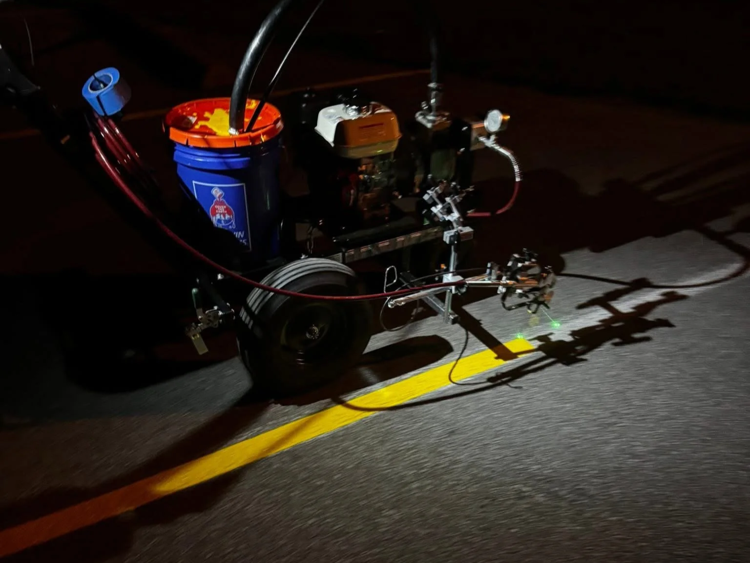 Automated machine with wheels and mechanical arms, illuminated by green lights, painted yellow line on dark pavement.