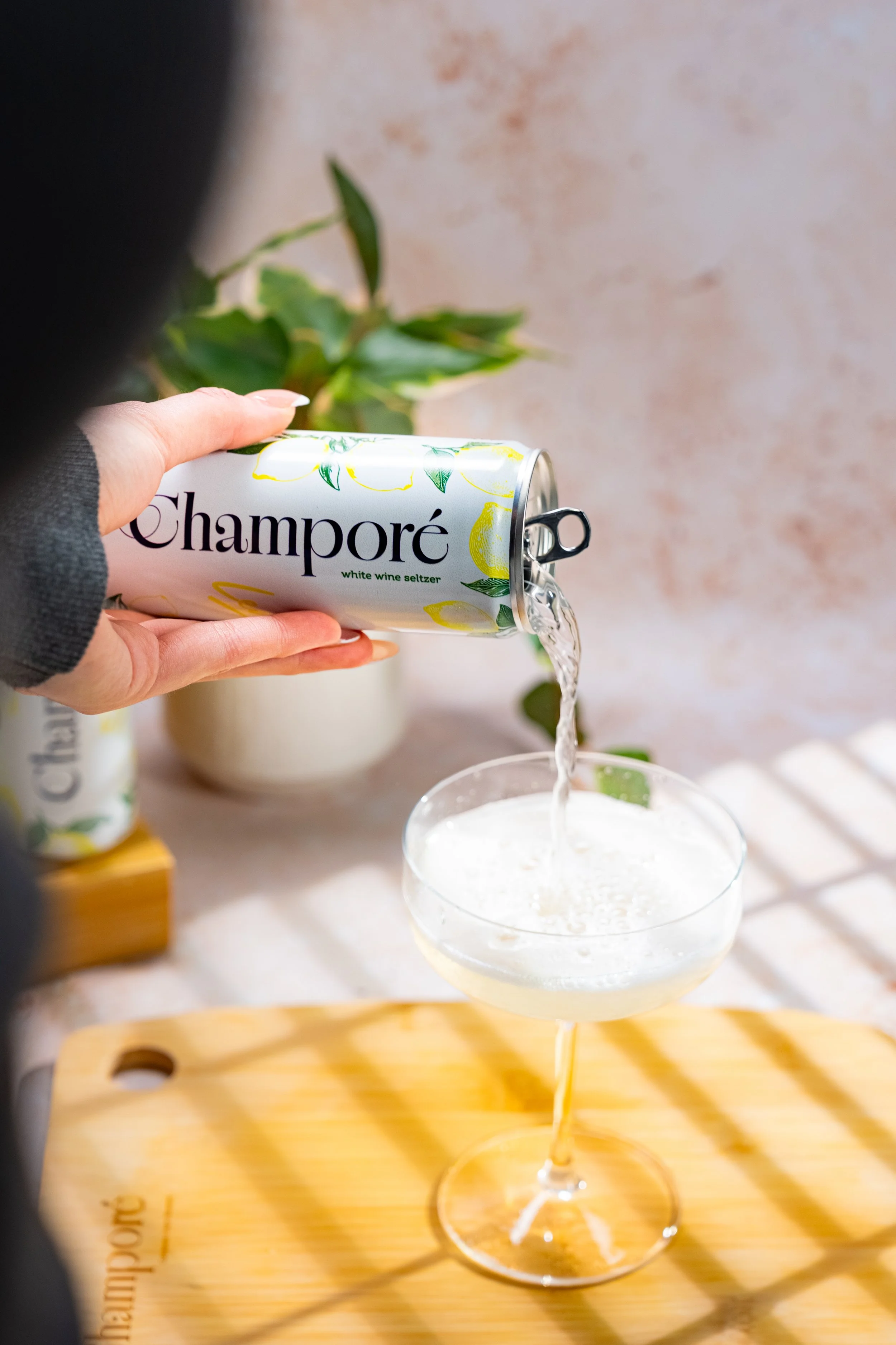 A hand holding a can labeled 'Champoré Lemon' pouring a sparkling white beverage into a cocktail glass. The scene is set on a wooden tray with a blurred background which includes a potted plant and a neutral-colored wall.