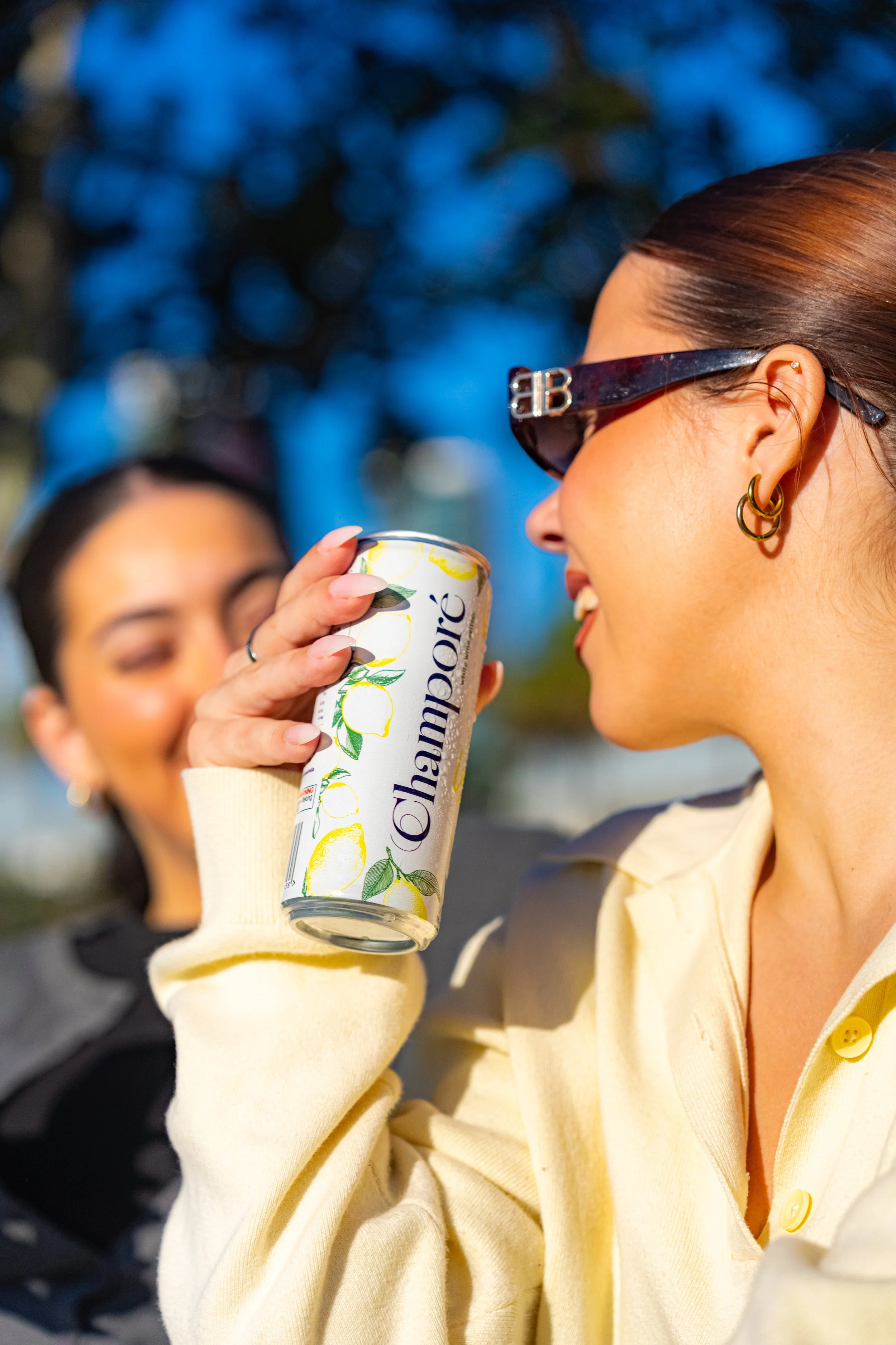 Close-up of a woman with brown hair, wearing large sunglasses and gold hoop earrings, holding a can of Champoré Lemon and smiling outdoors with another woman in the background.