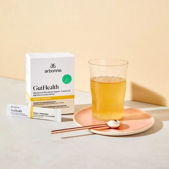 A box of Arbonne GutHealth supplement, a packet of Ginger flavored supplement, a glass of yellow liquid on a pink plate, with a spoon and chopsticks.