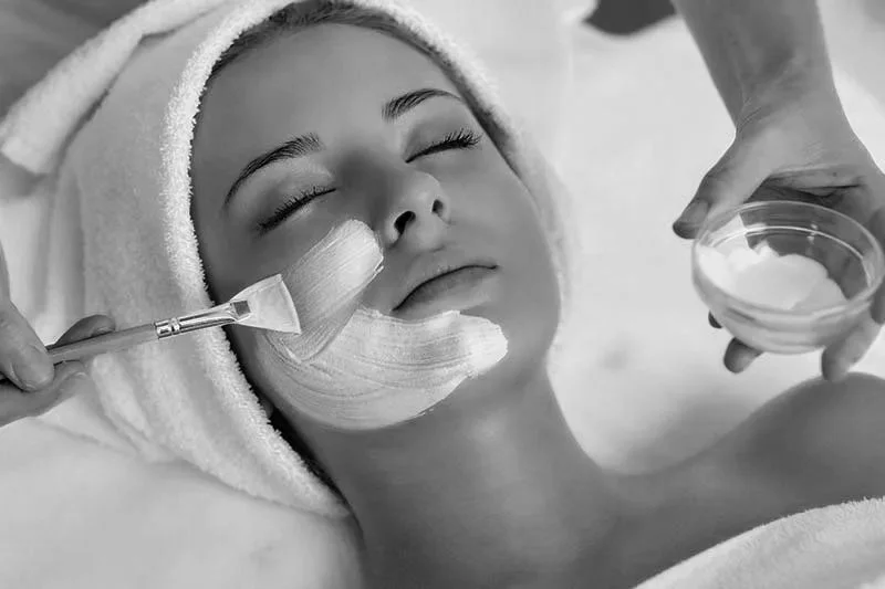 Woman with eyes closed receiving a facial treatment, with a towel wrapped around her head, and creams being applied with a brush and spatula