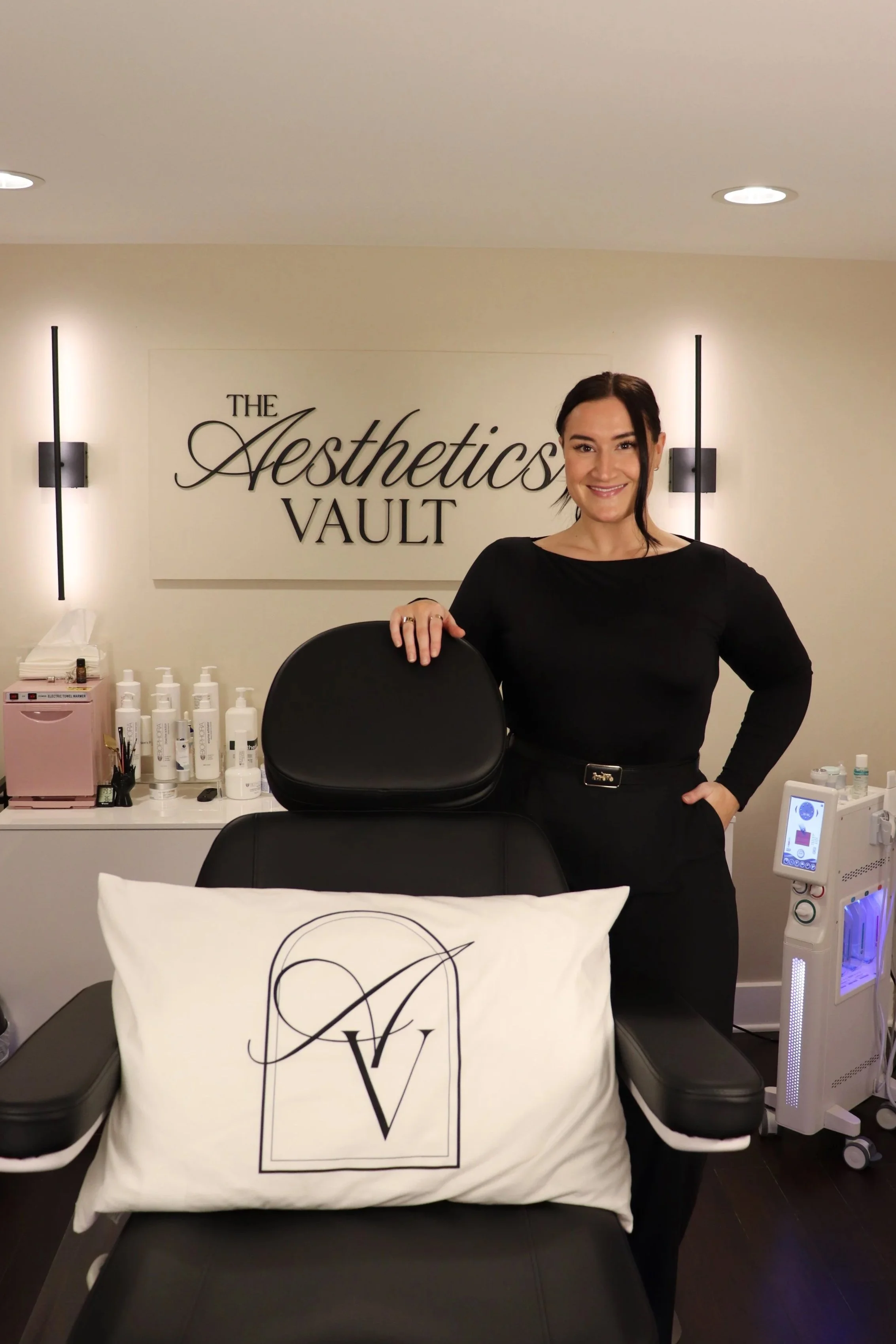 A woman dressed in black standing behind a treatment chair with a pillow marked with a logo, in a spa or clinic setting called 'The Aesthetics Vault'.