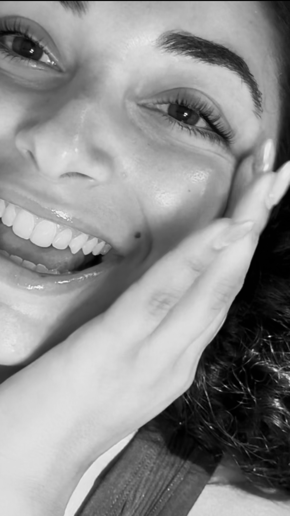 Close-up black and white photo of a smiling woman with dark hair, showing her teeth, with her hand gently touching her face.