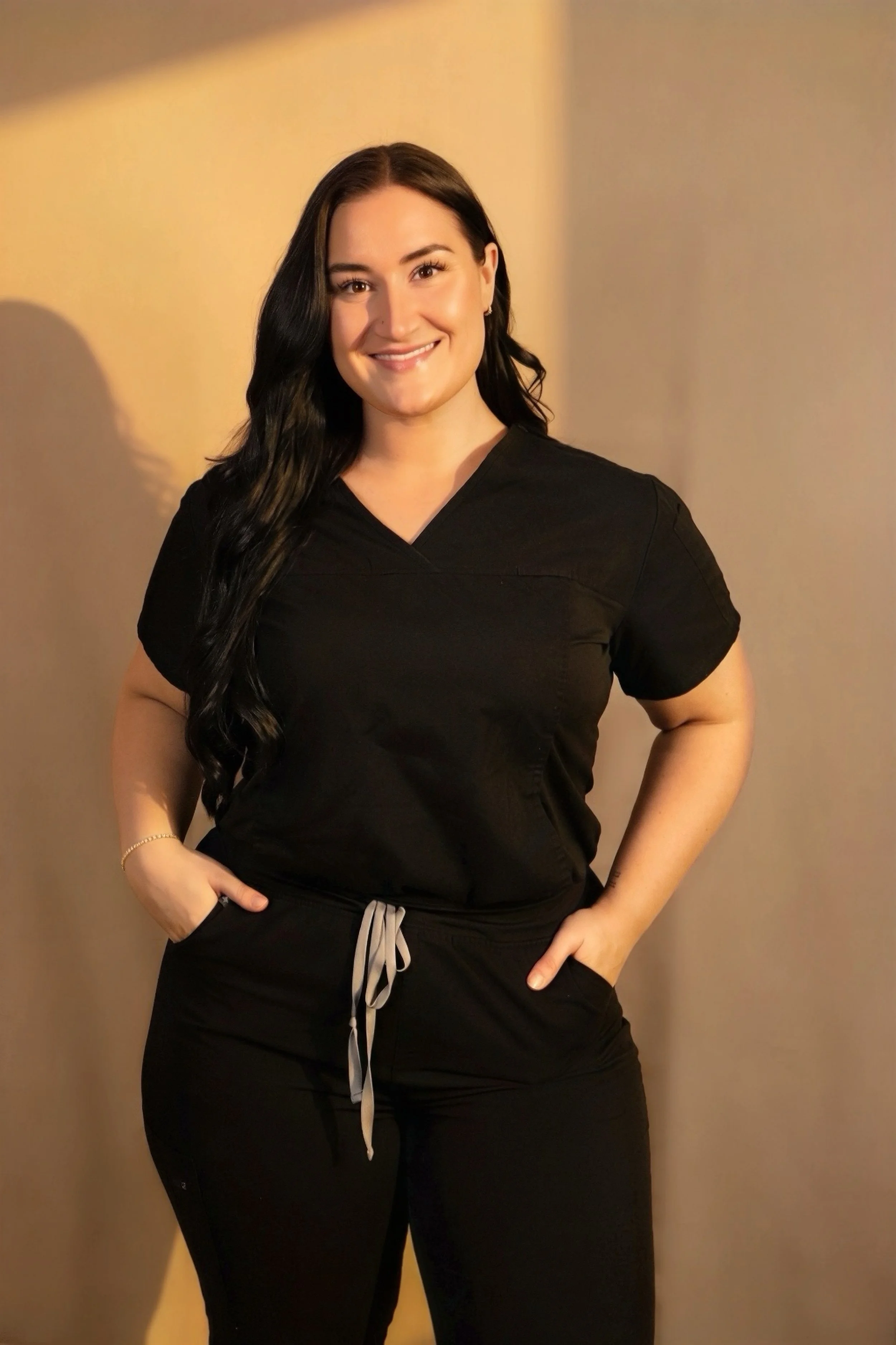 A smiling woman with long black hair wearing black medical scrubs and standing with her hands in her pockets, in front of a plain background.