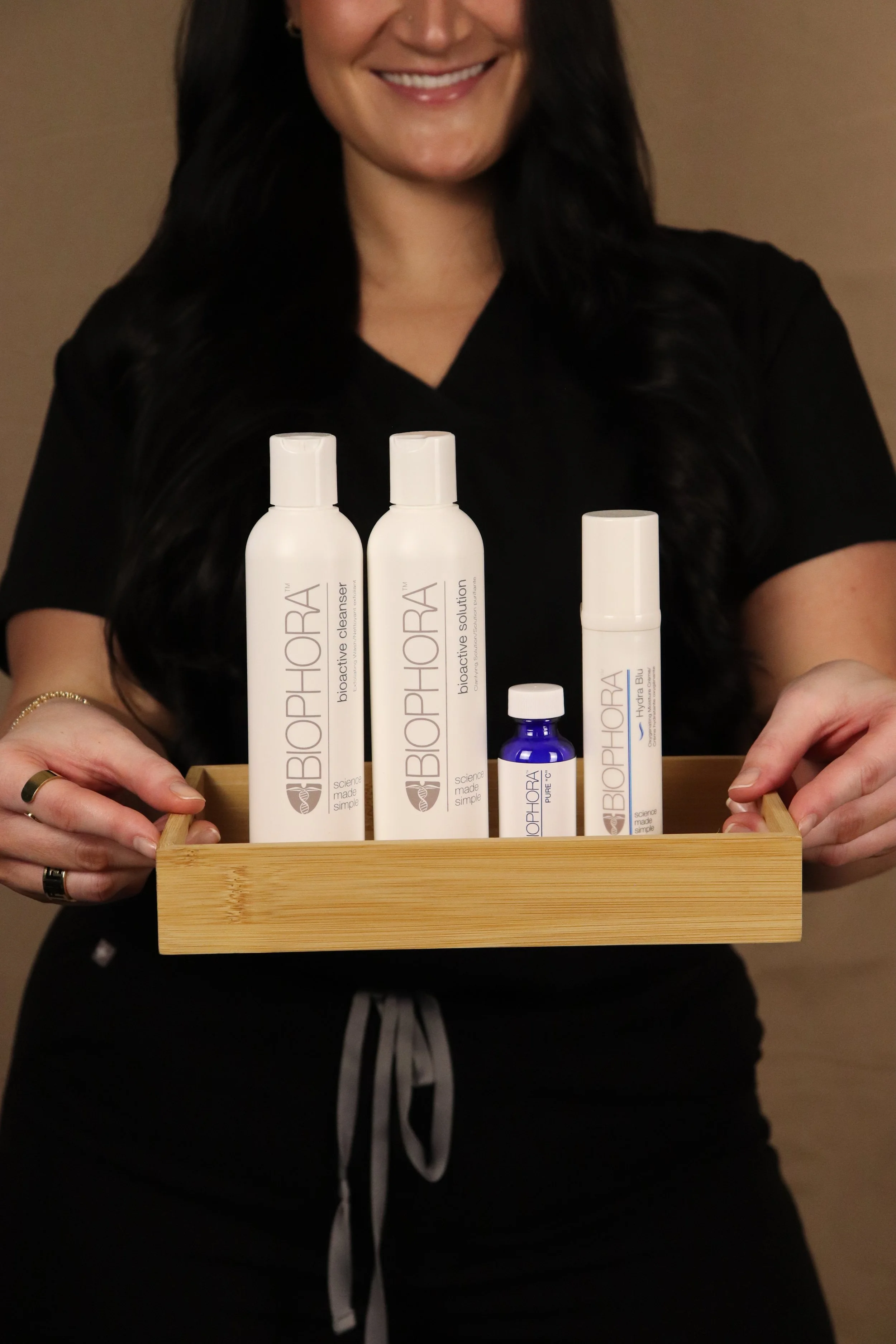 A woman holding a wooden tray containing various BIORPHORA skincare products, including two bottles of Bioactive Cleanser, a small bottle of Bioactive Solution, a larger bottle of Biohora Blue, and a small blue bottle, against a plain background.