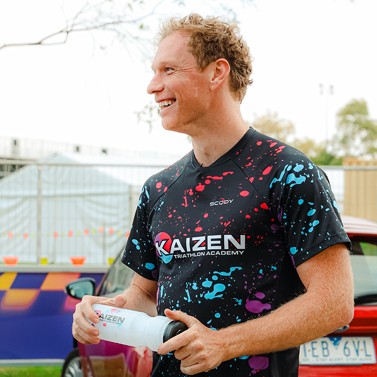 Smiling man with curly hair wearing a black triathlon shirt with colorful paint splatters and the logo 'KAIZEN Triathlon Academy', holding a white water bottle, outdoors with cars and a fence in the background.
