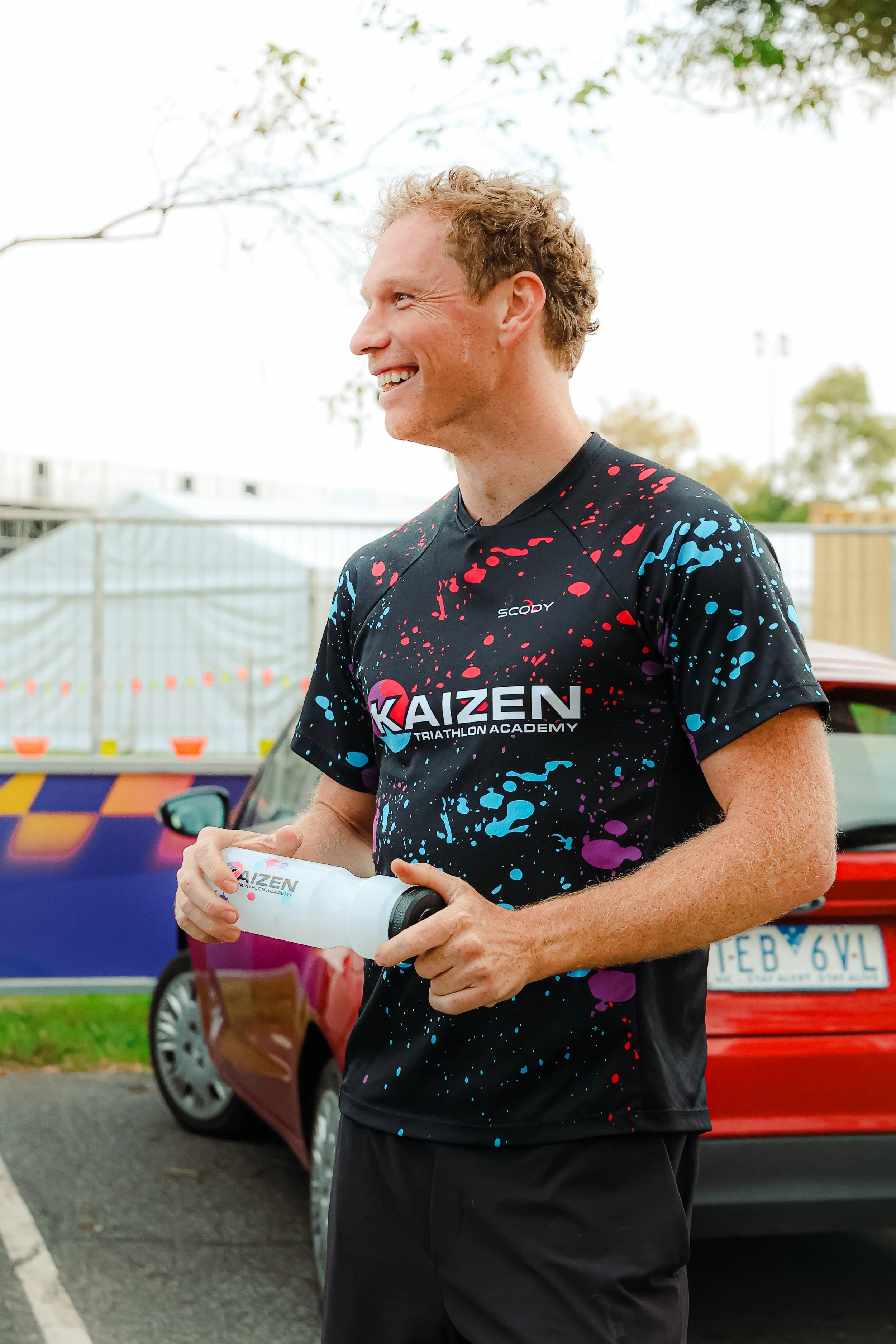 A smiling man in a black Kaizen Triathlon Academy t-shirt holding a water bottle, standing outdoors near parked cars.