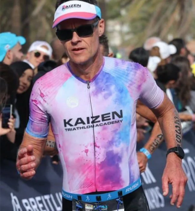 A male triathlete running in a race, wearing a colorful, tie-dye patterned cycling jersey that says 'KAIZEN TRIATHLON ACADEMY,' black sunglasses, a white and pink cap, and a black watch on his left wrist, with spectators in the background.