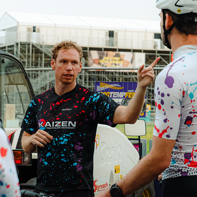 A man with curly hair wearing a black Kaizen Triathlon Academy shirt is talking to a cyclist with a helmet, both wearing colorful splatter-patterned jerseys, near a fuel station or pit stop.