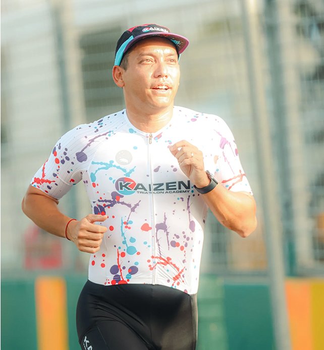 Man running outdoors, wearing a colorful cycling jersey, black shorts, and a cap, during daytime.