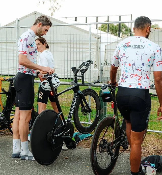 Three cyclists in colorful cycling jerseys preparing their bikes near a fence at an outdoor event.