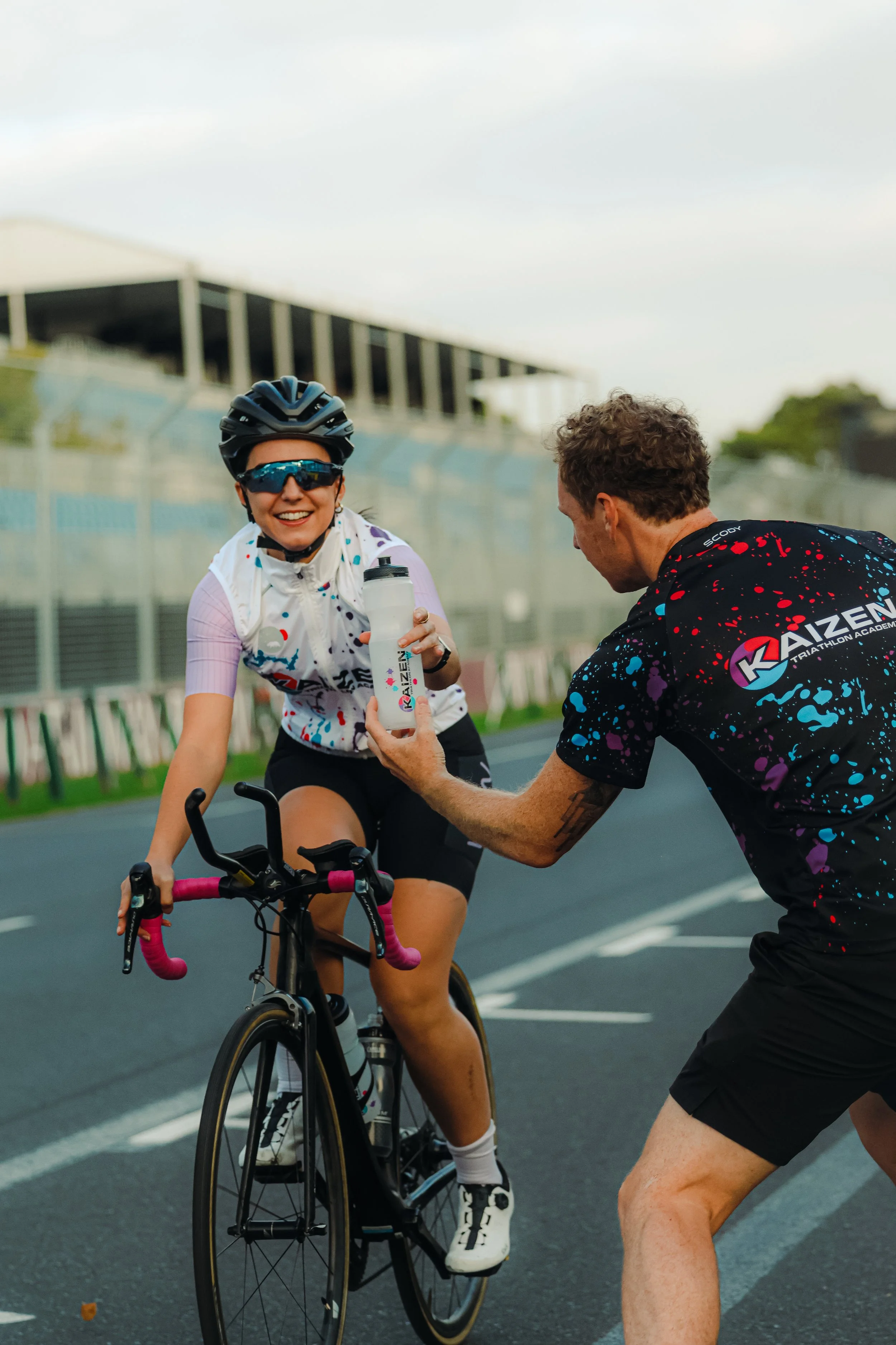 A woman on a bicycle receives a water bottle from a man during a cycling event on a race track.