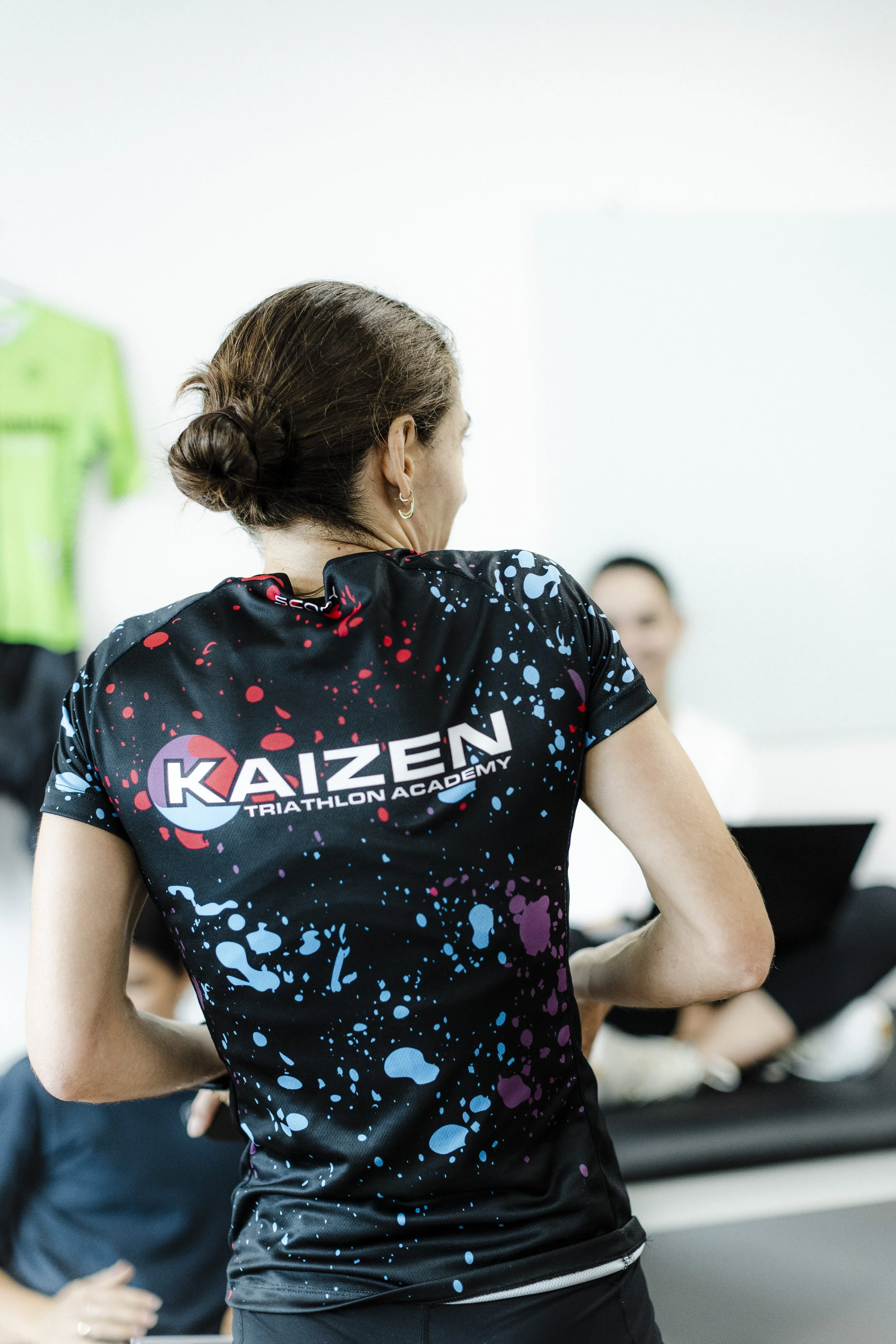 Back of woman wearing black athletic shirt with colorful splatters and 'KAIZEN TRIATHLON ACADEMY' logo, in indoor setting with other people and a laptop.