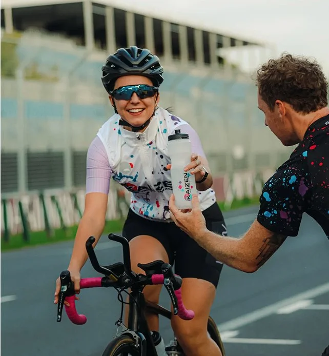A woman cyclist in a helmet and sunglasses smiling while receiving water from a man in a colorful biking jersey on a race track.