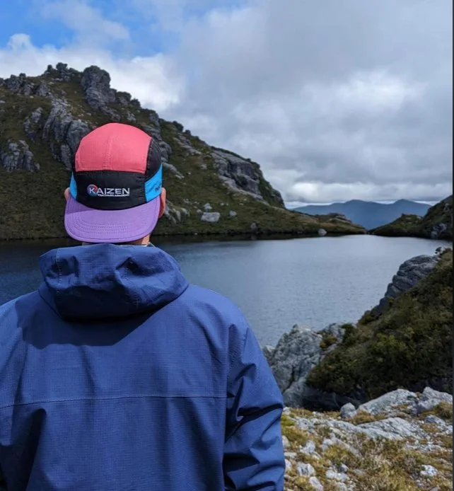 Person wearing a blue jacket and multicolored cap facing a lake surrounded by rocky hills under a cloudy sky.