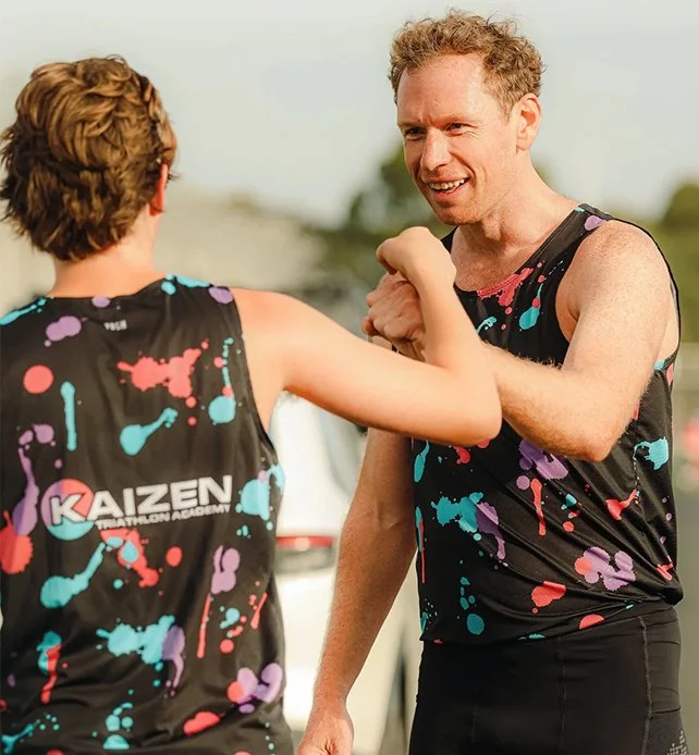 Two men in black athletic shirts with colorful splatter patterns, smiling and fist-bumping outdoors.