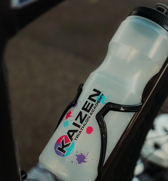 A white water bottle with a black cap, attached to a bicycle frame, featuring the Kazen Triathlon Academy logo with colorful paint splashes.