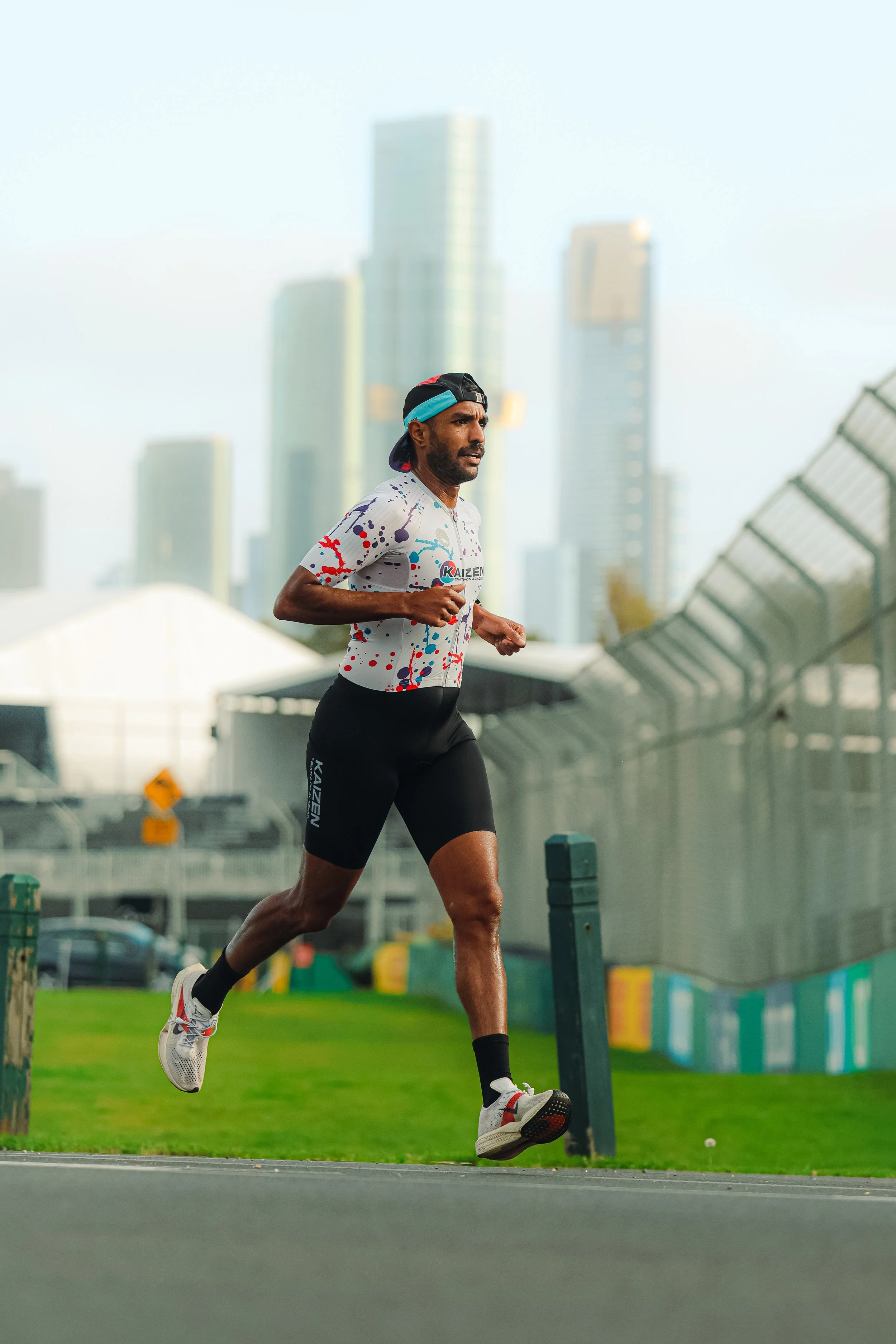 A man running outdoors on a city street with tall buildings in the background, wearing a colorful t-shirt, black shorts, a cap, and running shoes.