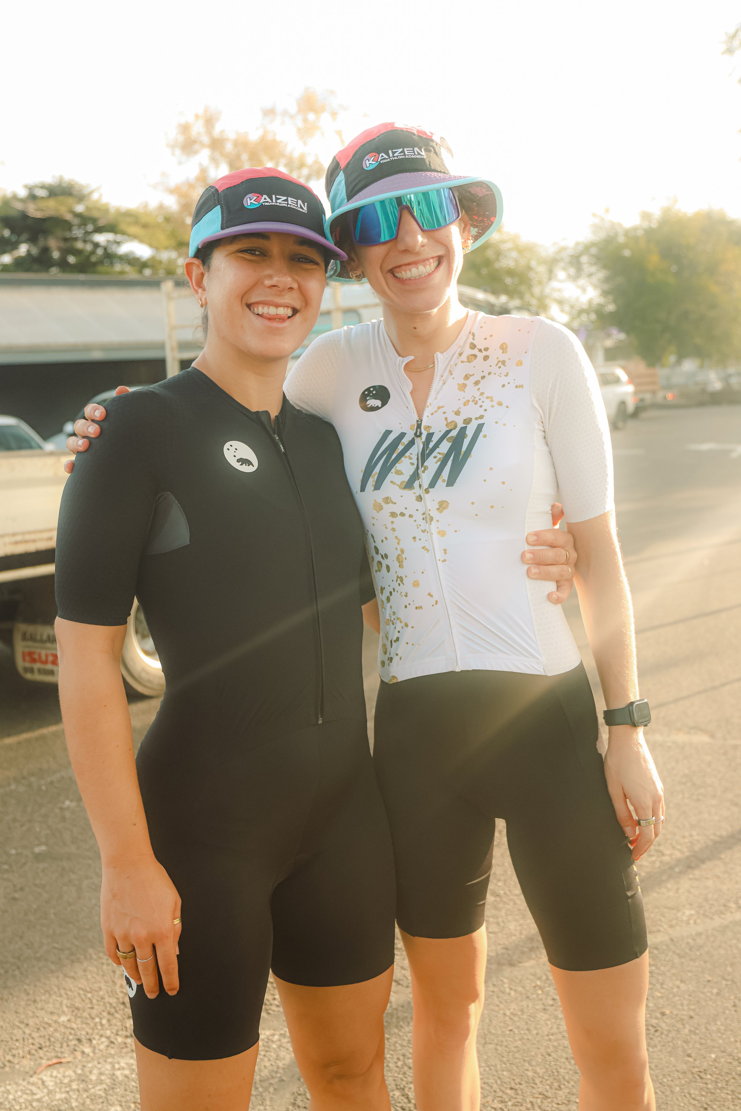 Two women in athletic clothing and caps standing outdoors, smiling, with one woman having her arm around the other's shoulder, during a sunny day.