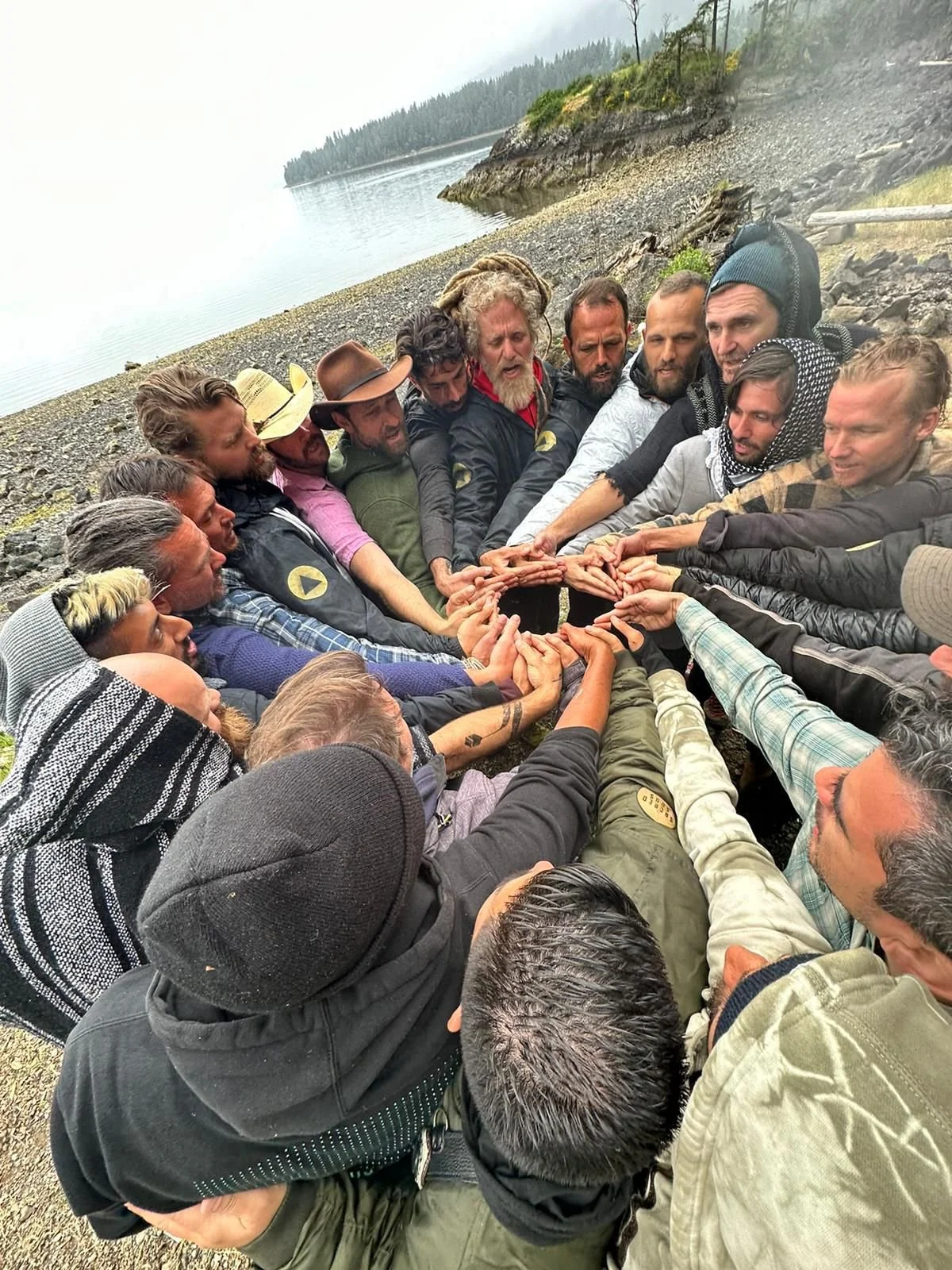 Group of people outdoors on a rocky beach, forming a circle with their hands overlapping in the center, near a lake with trees in the background.