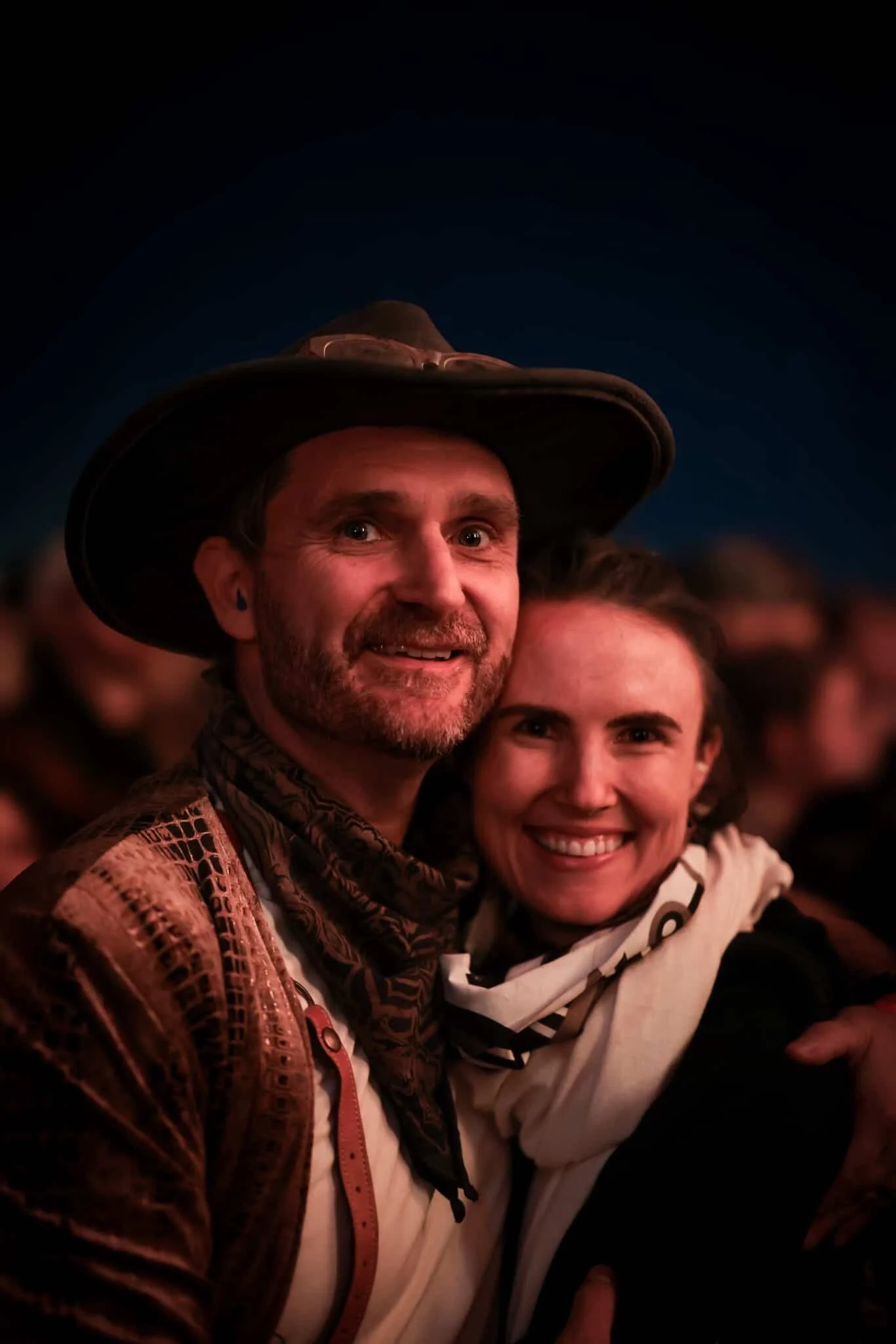Two smiling people, a man with a wide-brimmed hat and a woman, are close together at an event, with a blurred crowd in the background.