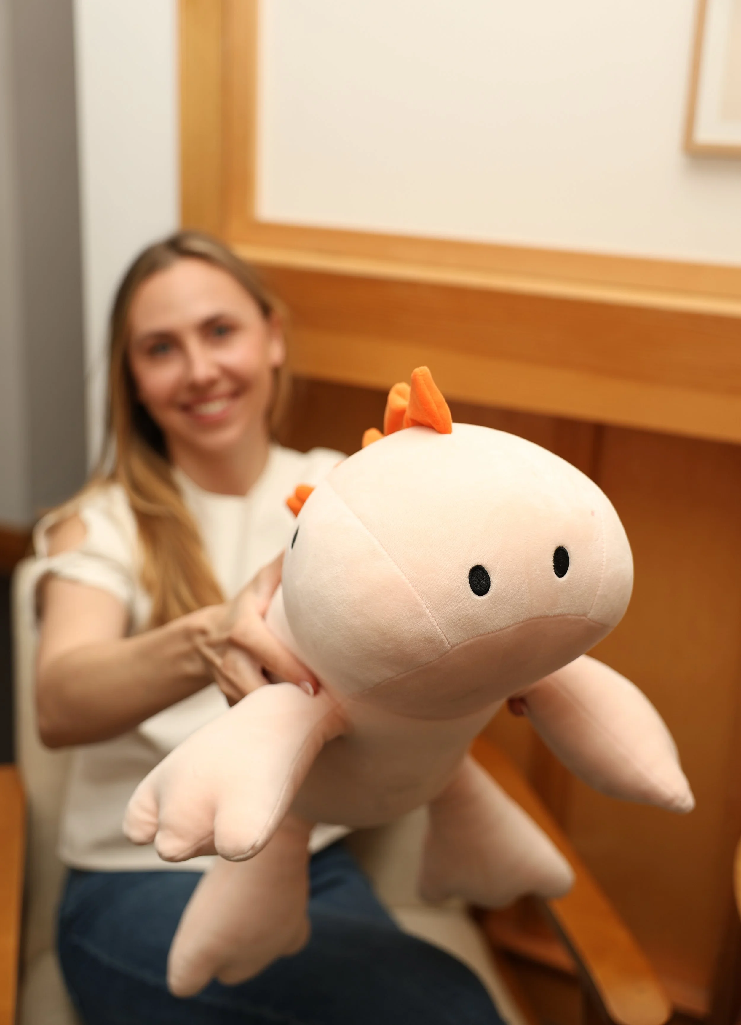 A woman smiling and sitting indoors, holding a large plush toy of a cartoon dinosaur with orange spikes on its back.