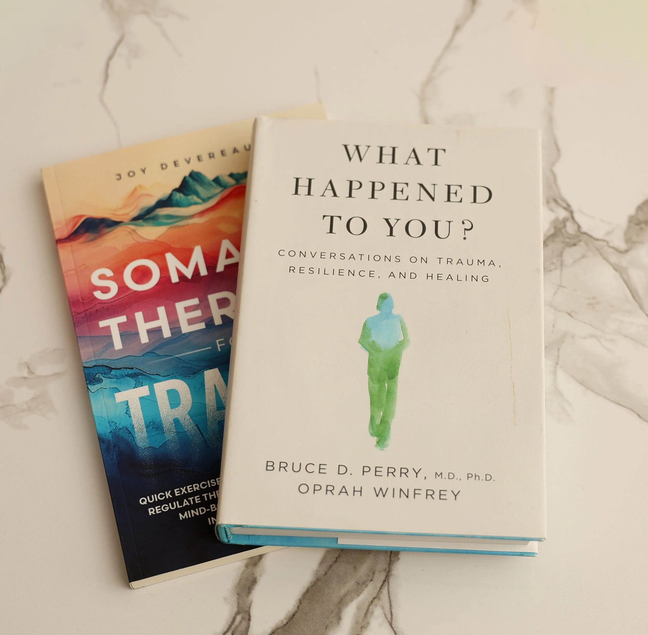 Two books, one titled 'What Happened to You?' and another with a colorful cover about somatic therapy, placed on a white marble surface.