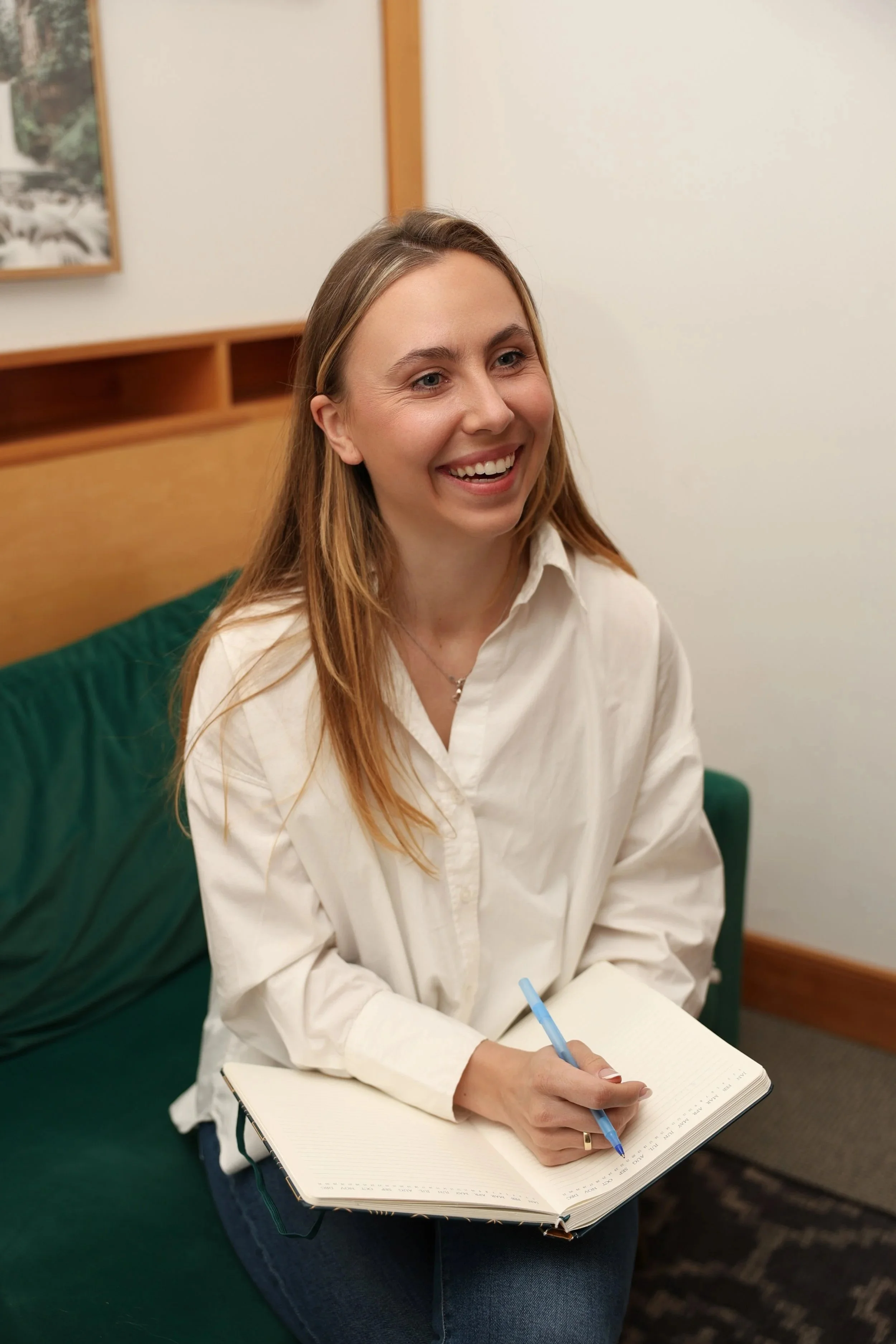 A young woman with long blonde hair, wearing a white button-up shirt, sitting on a green sofa in an office or waiting room, smiling, holding a blue pen, and writing in a notebook.